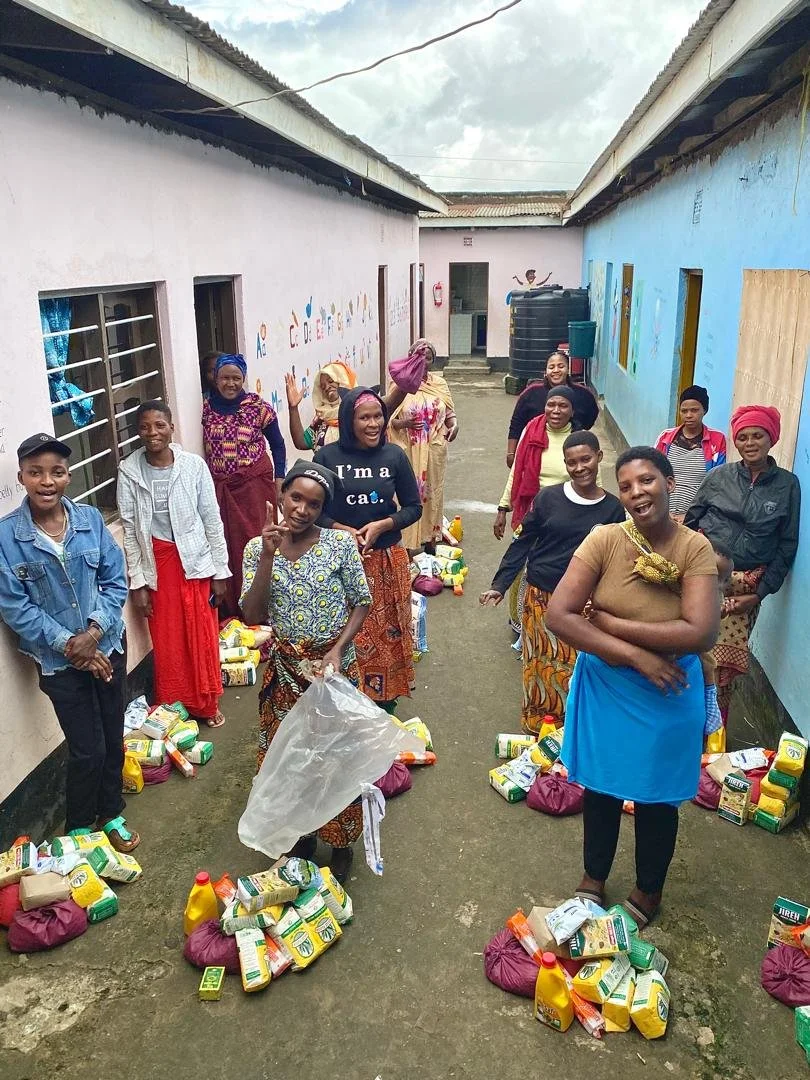 Group of women and children standing in a narrow outdoor space between two colorful buildings, with charity supplies and food packages on the ground, smiles, and peace signs.