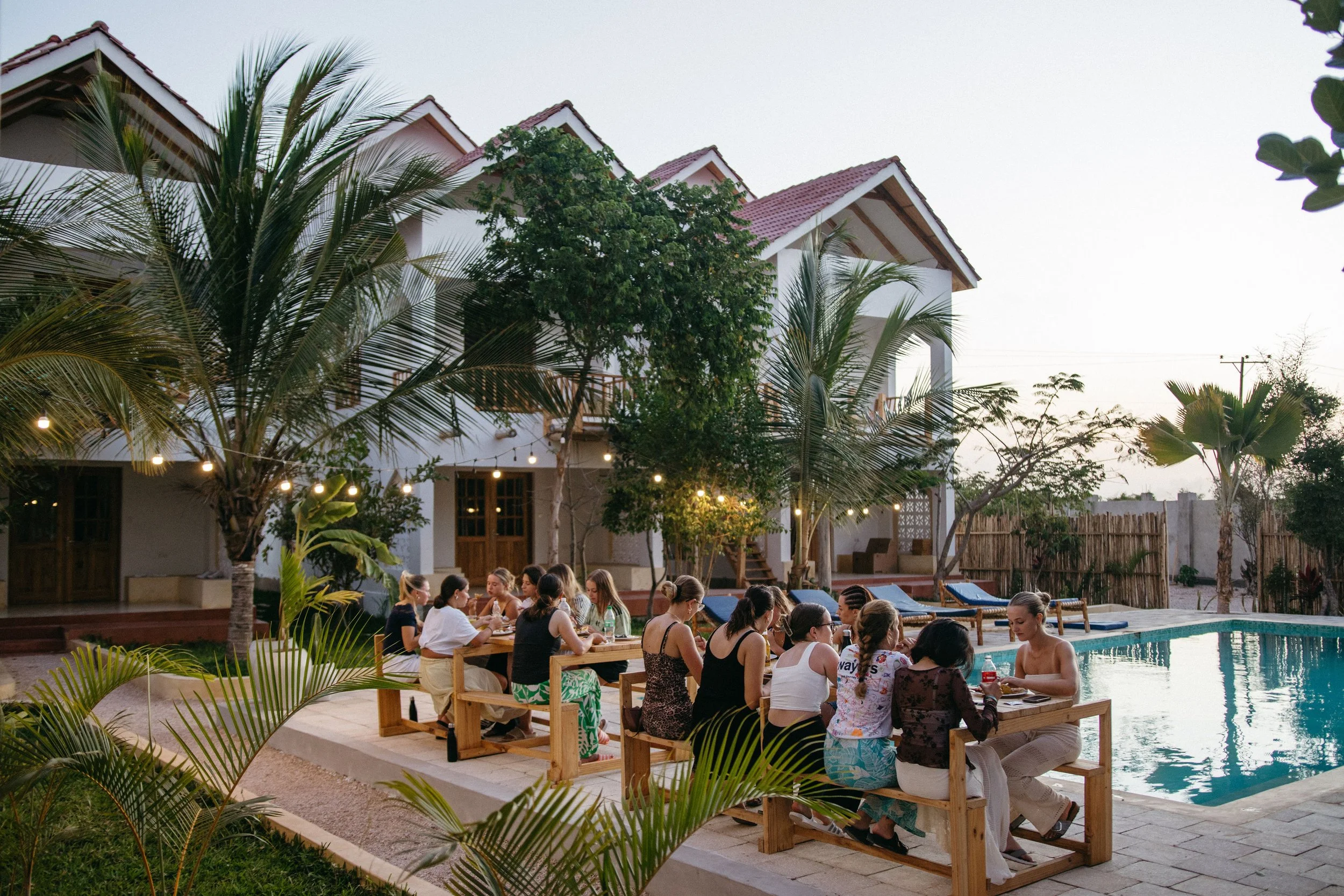 People sitting at tables by a swimming pool in a backyard, with a house, palm trees, and string lights in the background during evening.
