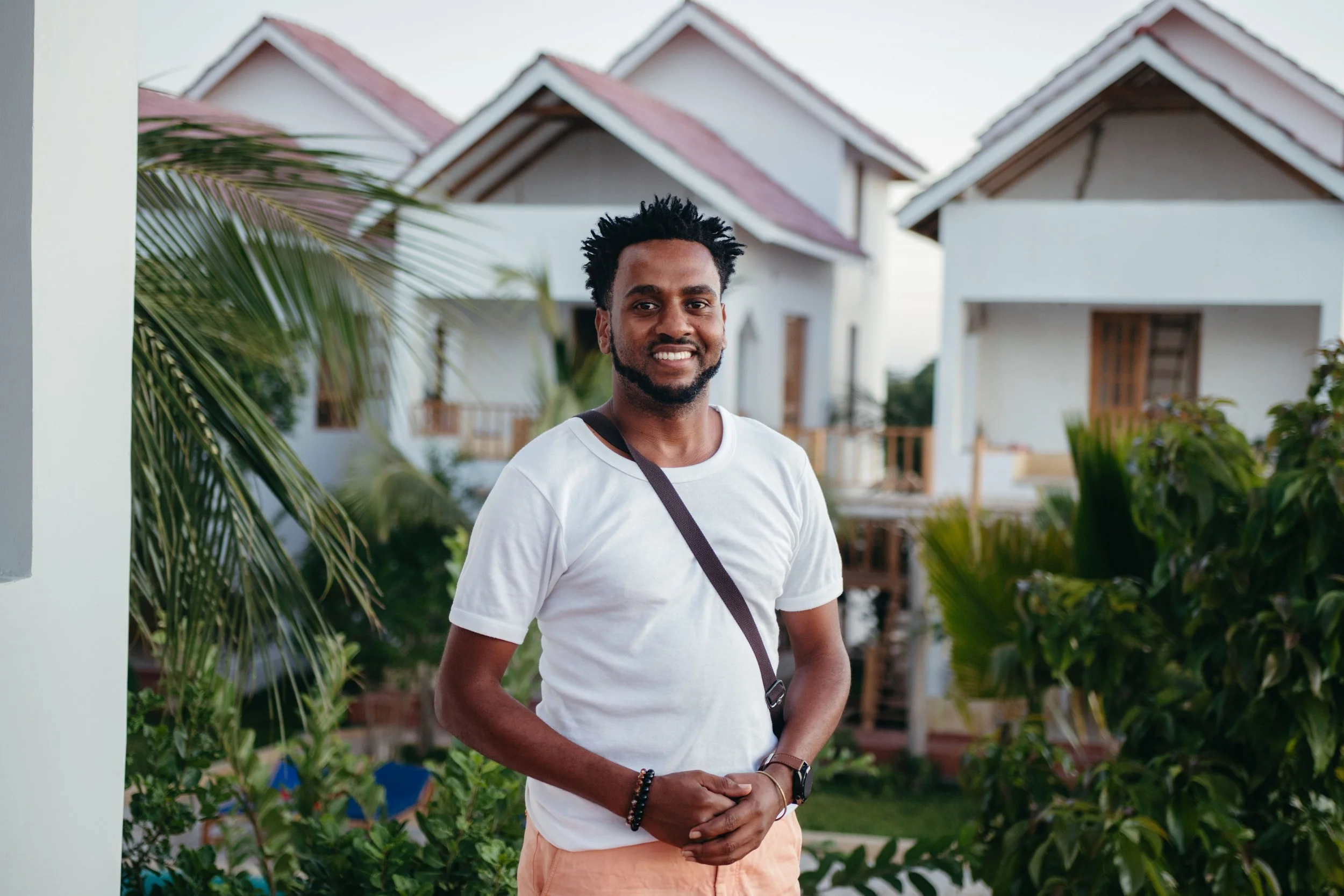 A man with dark skin, black hair, and a beard, smiling while standing outdoors near a white building with multiple houses in the background. He is wearing a white t-shirt, pink shorts, a black wristwatch, and black bracelets, with a black strap across his chest, likely from a bag or camera.