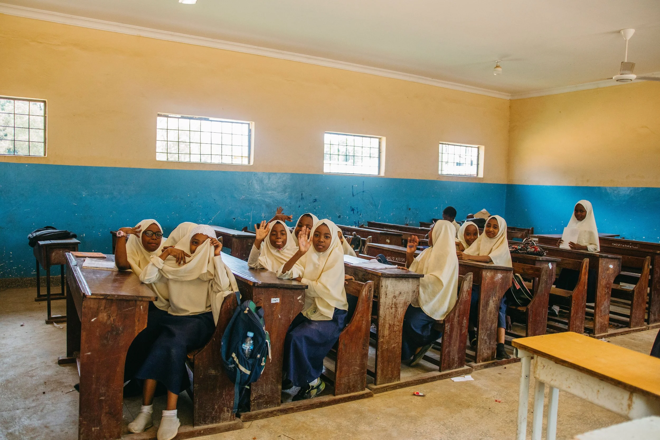 A classroom filled with young girls wearing cream-colored headscarves and uniforms, sitting at wooden desks and raising their hands. The classroom has yellow and blue walls with four small windows near the ceiling and a ceiling fan.