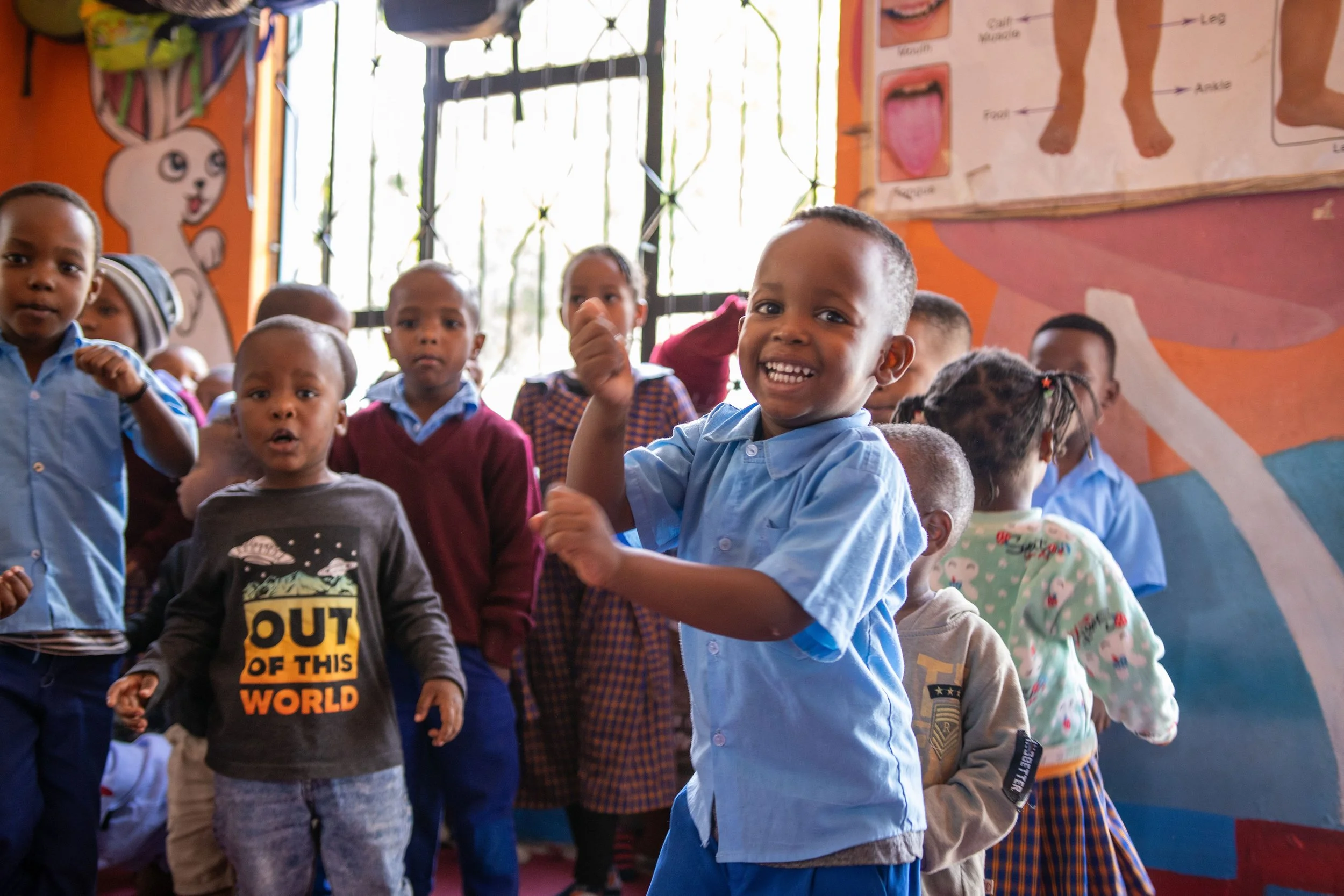 A group of young children in a classroom, some smiling and looking at the camera. They are wearing casual clothing, with some in school uniforms. The classroom has colorful walls with drawings and educational posters.