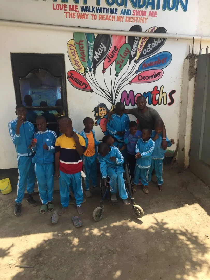 A group of children and a young adult standing in front of a colorful mural with balloons representing the months of the year on a school wall. The children are wearing blue sports uniforms, and one child in a wheelchair is present. The mural is titled 'Maths' and has a motivational message at the top.