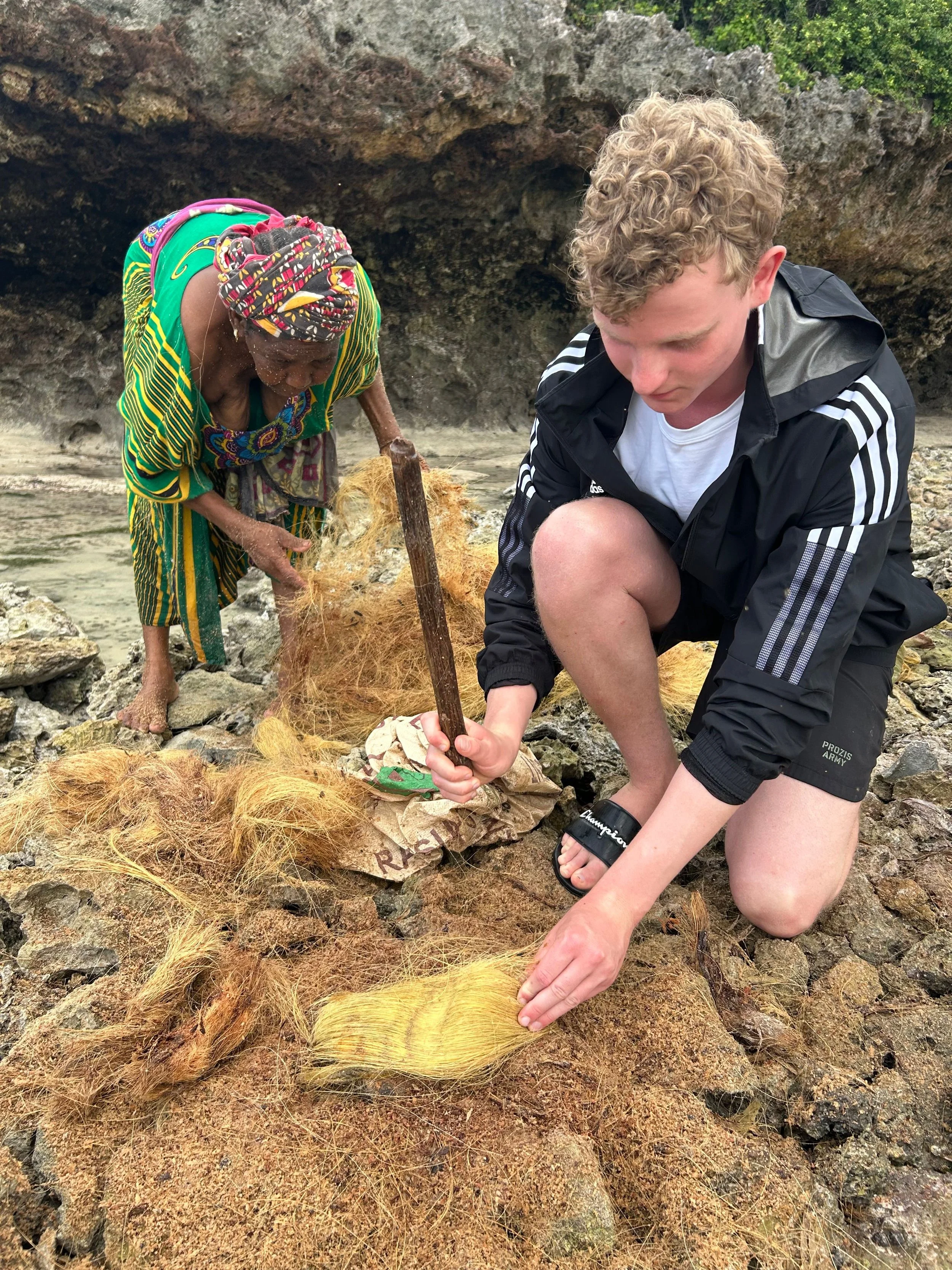 A woman and a man on a rocky beach handling a large bundle of yellow rope and a piece of cardboard with writing on it. The woman is wearing colorful clothing and a headscarf, while the man is dressed in a black jacket and shorts. They are kneeling on the ground surrounded by rocks and sand.