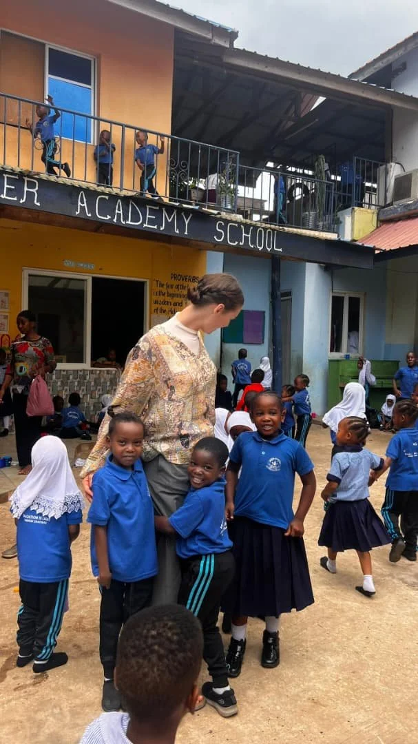 A group of children in blue school uniforms smiling with a teacher outside a school building labeled 'Teacher Academy School'. Some children are playing on the ground and others are standing near the teacher, while more children can be seen on a balcony above.