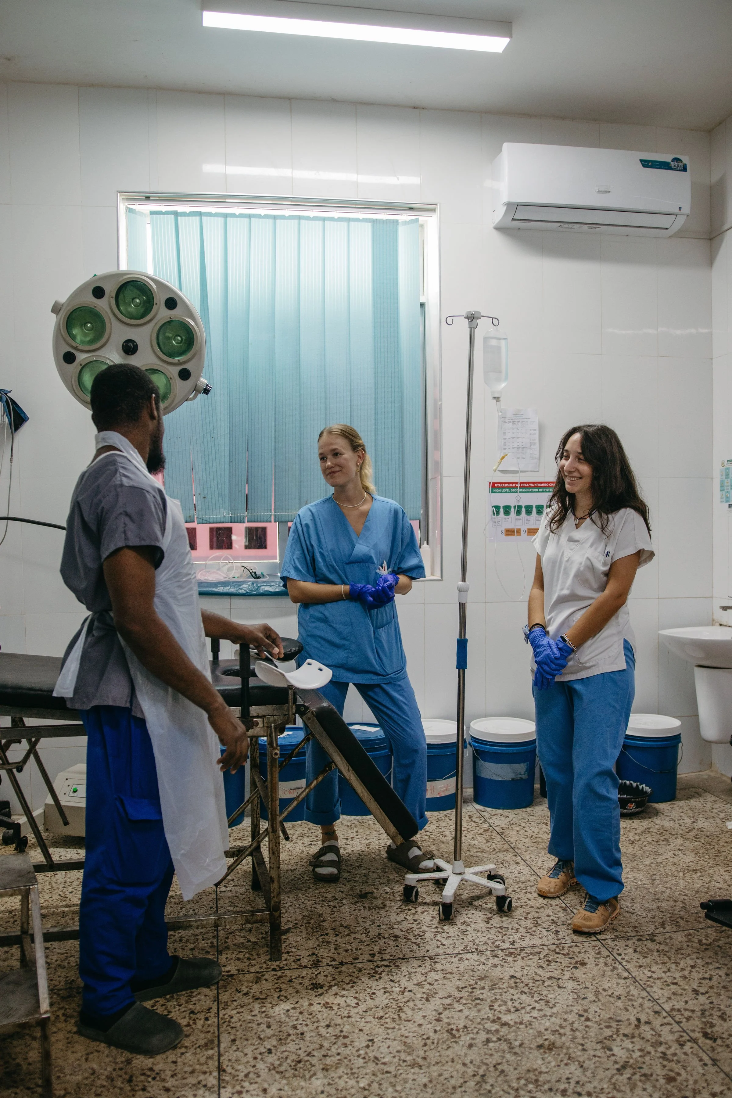 Medical professionals in scrubs and gloves in a hospital room, two women smiling, one man with a beard and dark hair, hospital equipment including an operating light, IV stand, and an anesthesia machine, with a window and blue blinds in the background.