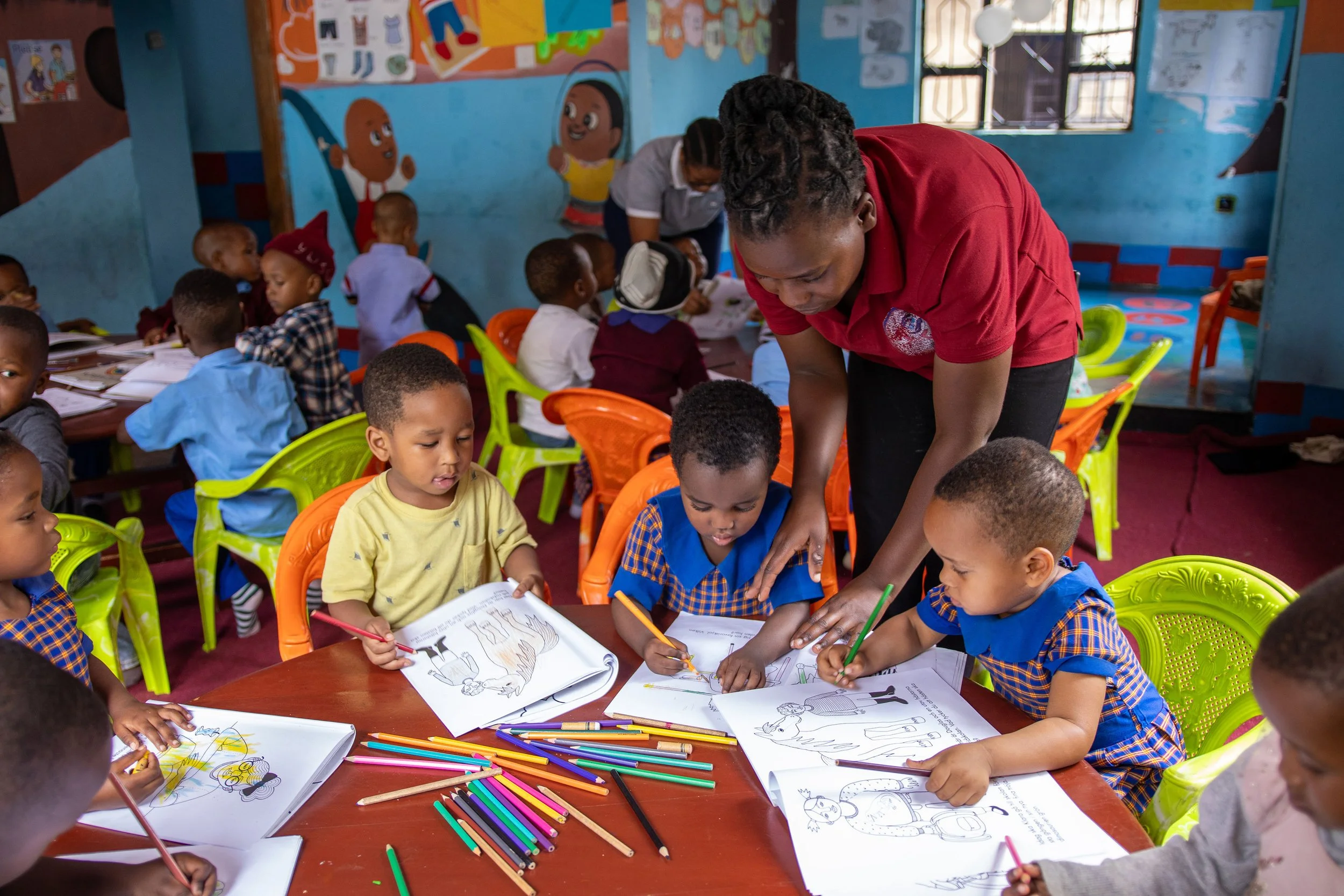 Kids in colorful uniforms participating in a classroom art activity with an adult teacher. The room has vibrant murals on the walls and art supplies like colored pencils on the table.