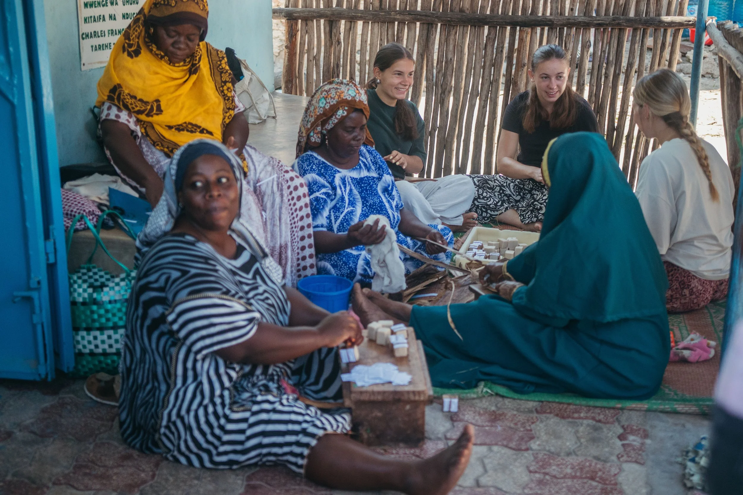 A group of women and girls sitting and kneeling on the ground under a makeshift bamboo roof, engaging in a health or community activity, with some exchanging small items or medication, surrounded by a simple rustic environment.