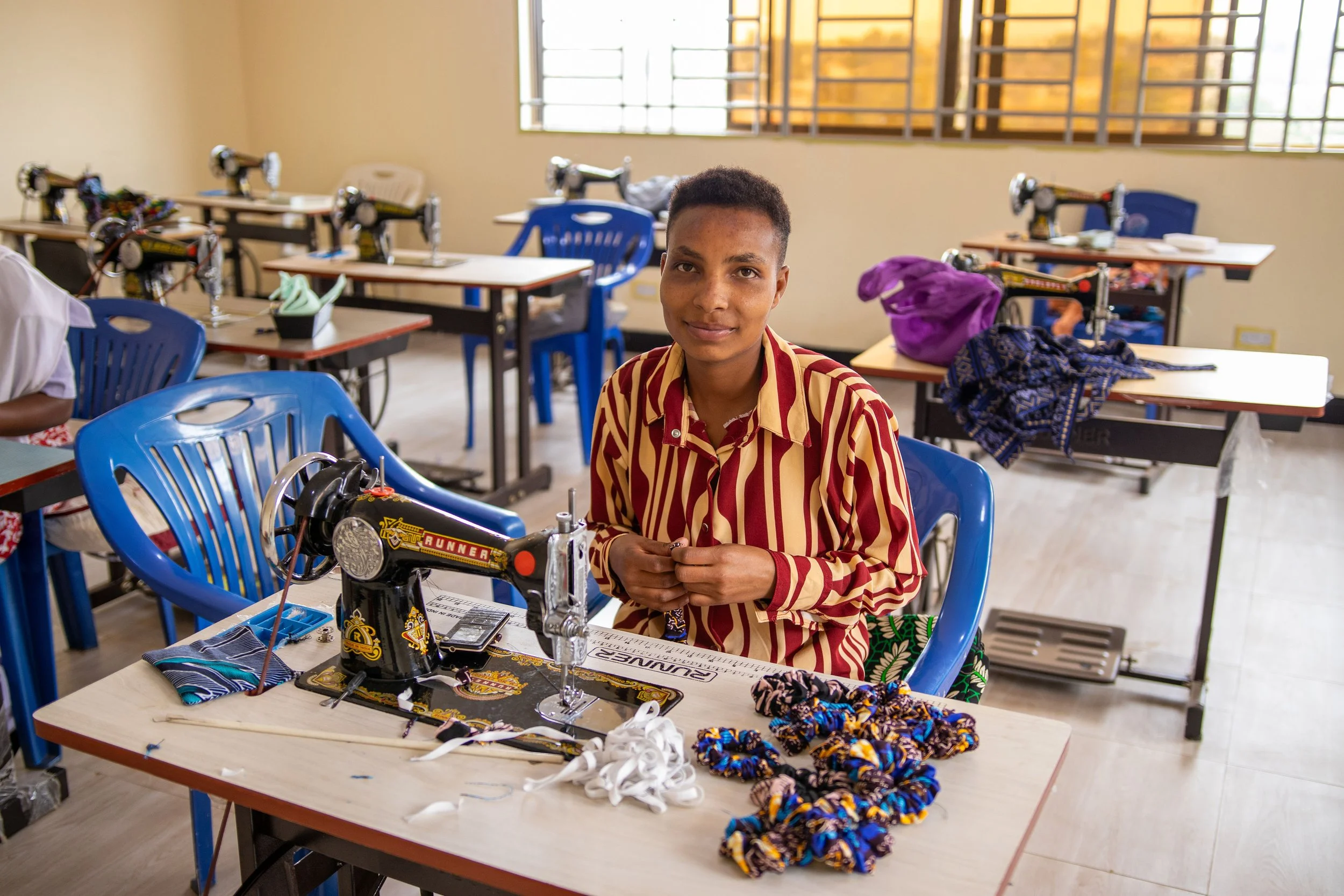A young boy sitting at a desk with a sewing machine in a classroom. The desk has various fabric scraps and sewing supplies. Other sewing machines and desks are visible in the background near windows.