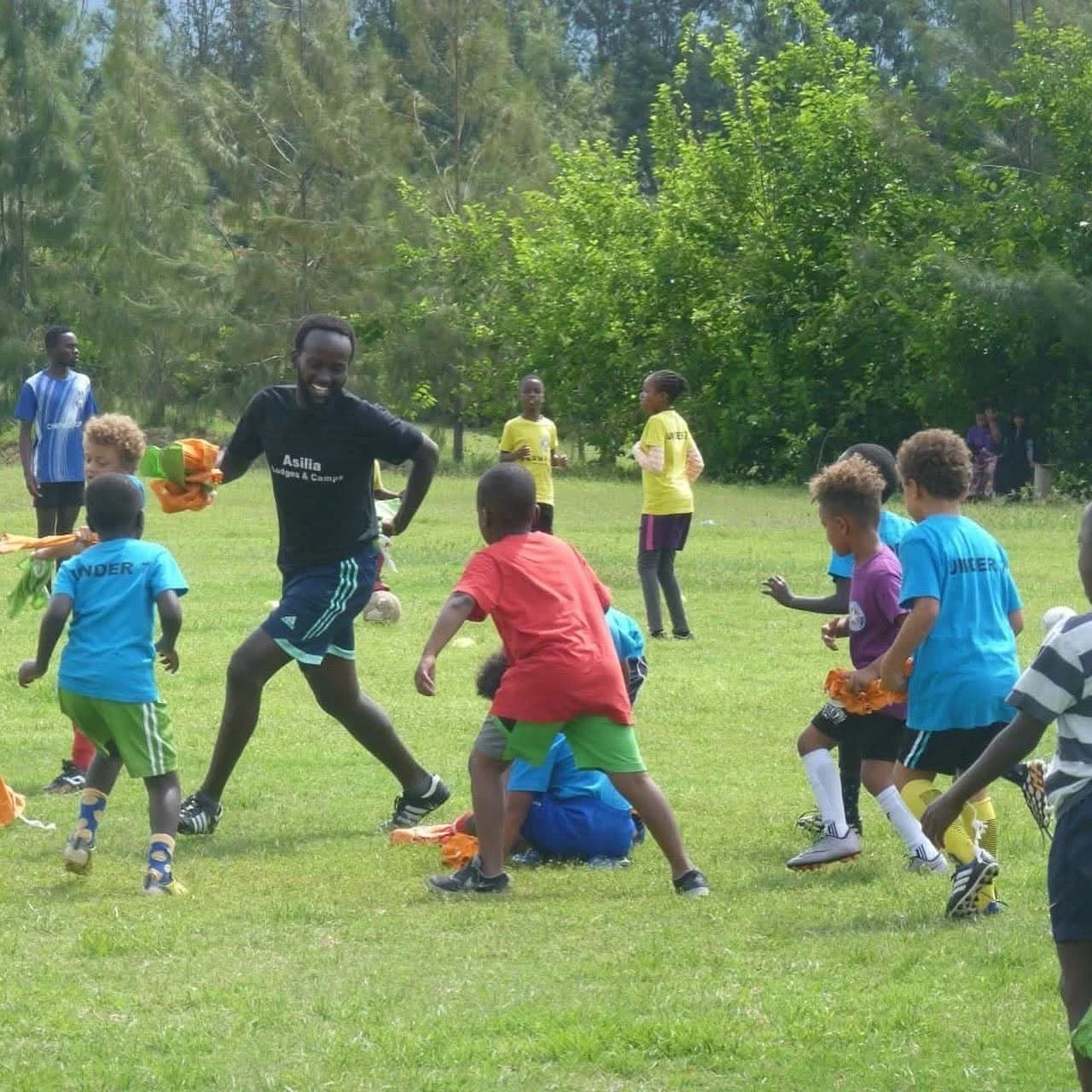Children playing soccer outdoors on a grassy field with trees in the background, while an adult coach runs among them.
