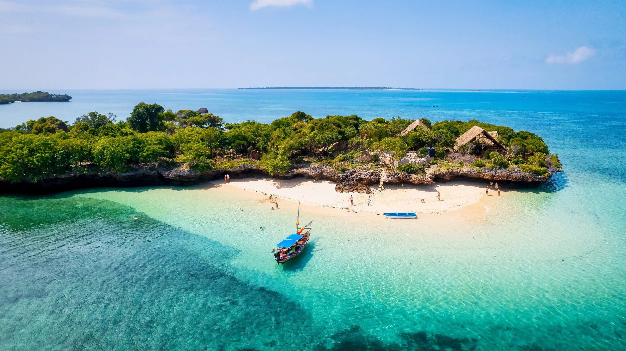 A tropical island with white sandy beach, surrounded by turquoise water, lush green trees, and thatched-roof huts, with a boat and people enjoying the beach and water.