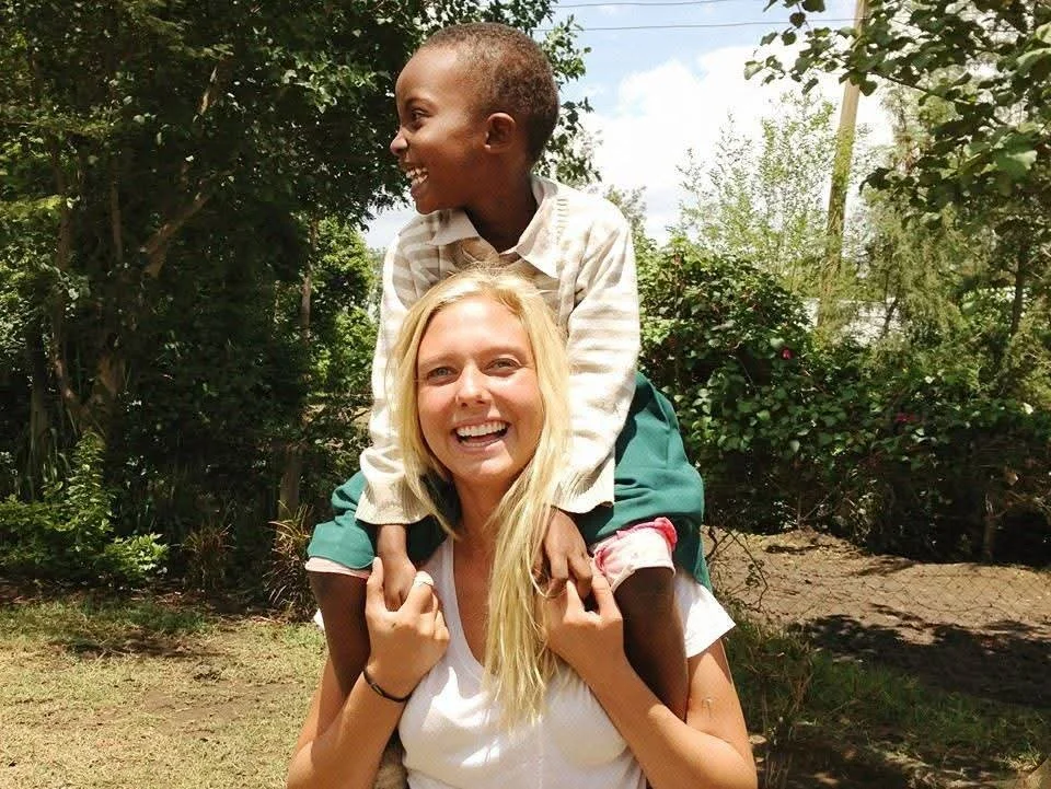 A smiling young woman with long blonde hair giving a piggyback ride to a boy with short hair, outdoors in a garden with trees and bushes.