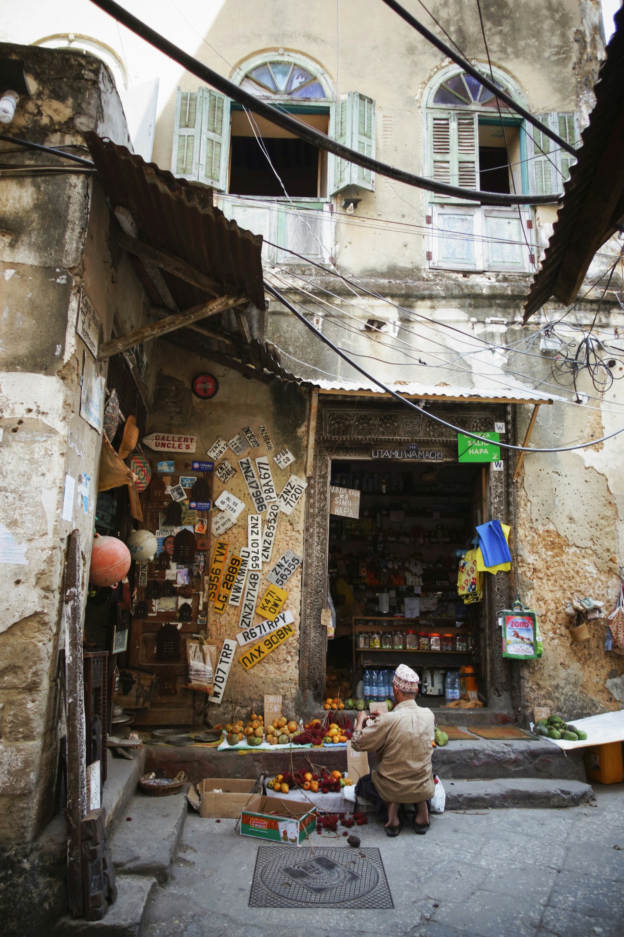 A street vendor sells tropical fruits such as oranges, pomegranates, and dragon fruits outside a small shop in a weathered building with boarded-up windows and a cluttered exterior. A man is sitting on a stool in front of the stall with a bag.