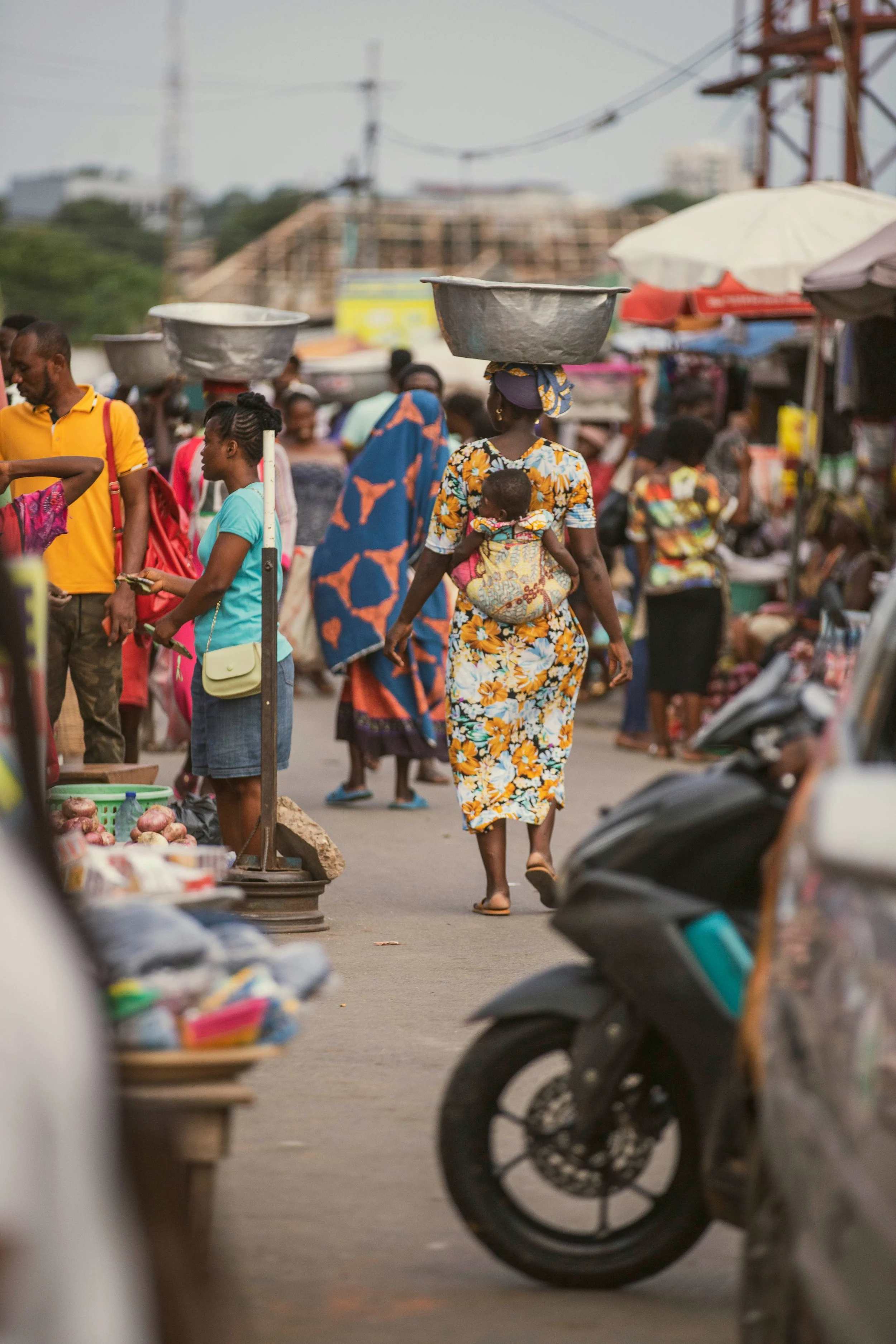 A woman walking in a busy outdoor market carrying a large metal basin on her head, with a baby strapped to her back. The market is filled with people, stalls, umbrellas, and scooters, in an urban setting.