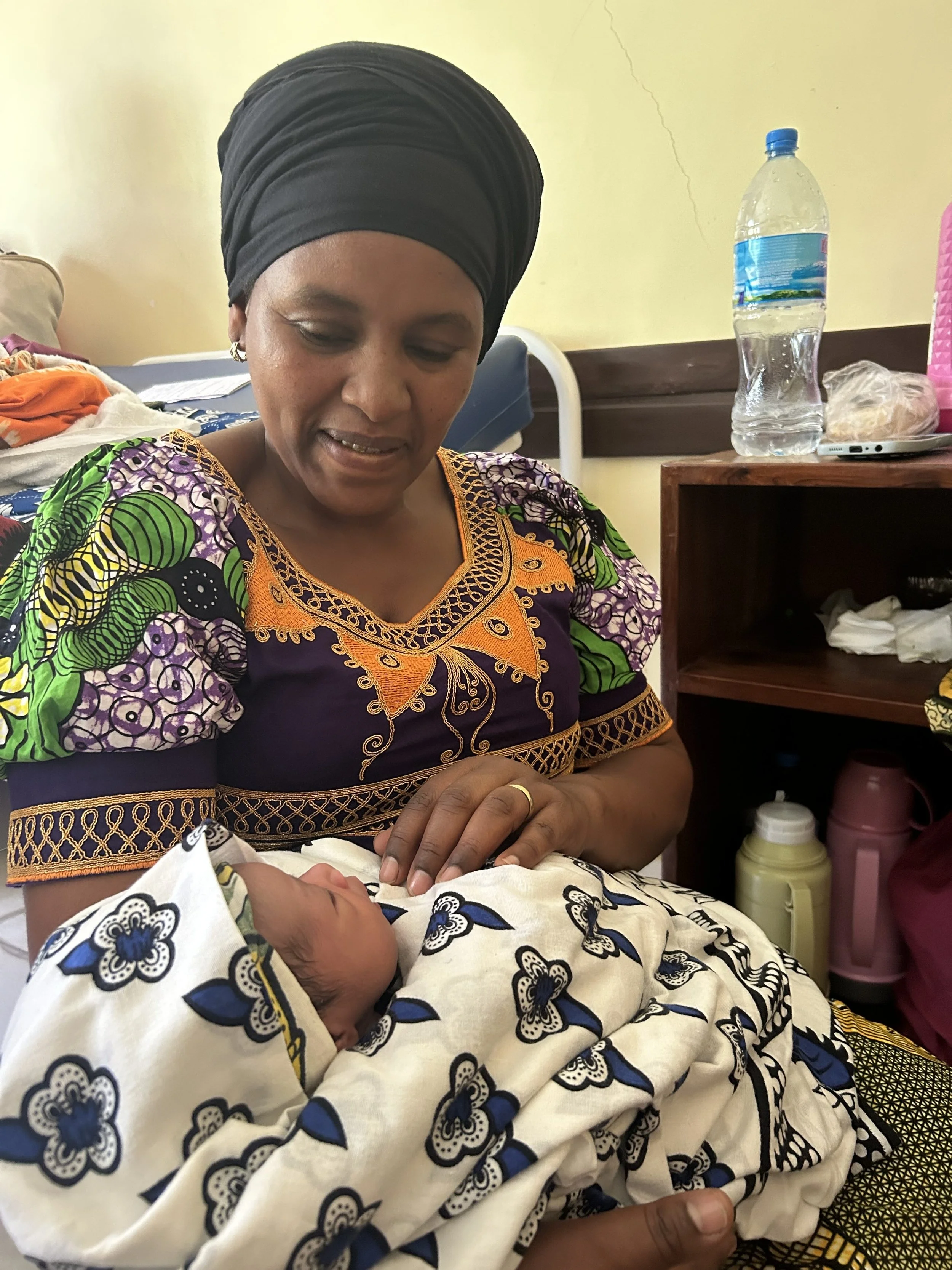 A woman wearing a colorful, traditional African dress and headwrap is looking at a newborn baby wrapped in patterned cloth. The woman is smiling and touching the baby's hand in a hospital room.