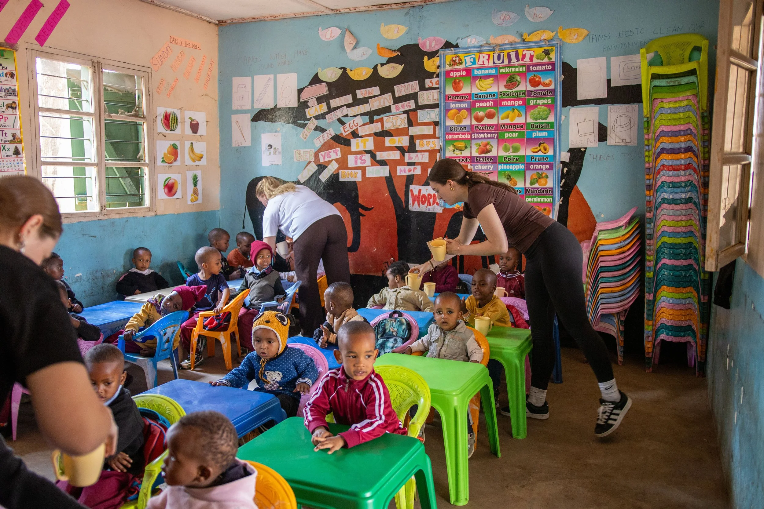 Children sitting at colorful tables in a classroom, with teachers distributing drinks. The classroom walls are decorated with educational posters and drawings, including a fruit chart and sound words.