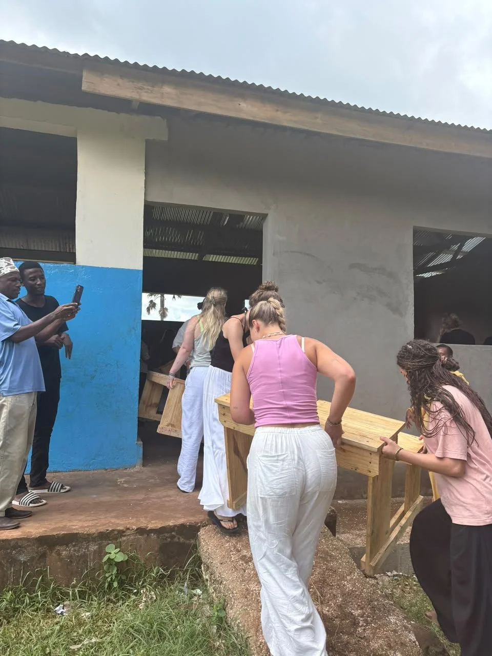 Group of women carrying a wooden table up a small ramp outside a building with concrete walls and a corrugated metal roof, while others observe and take photos.