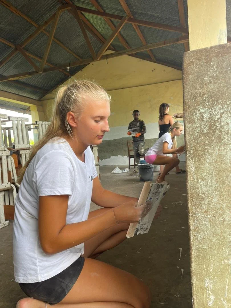 A young woman with long blond hair in a ponytail is kneeling on the ground, holding a palette and painting on a wall. Three other women are painting a wall in the background inside a partially constructed or renovated building with a corrugated metal roof.