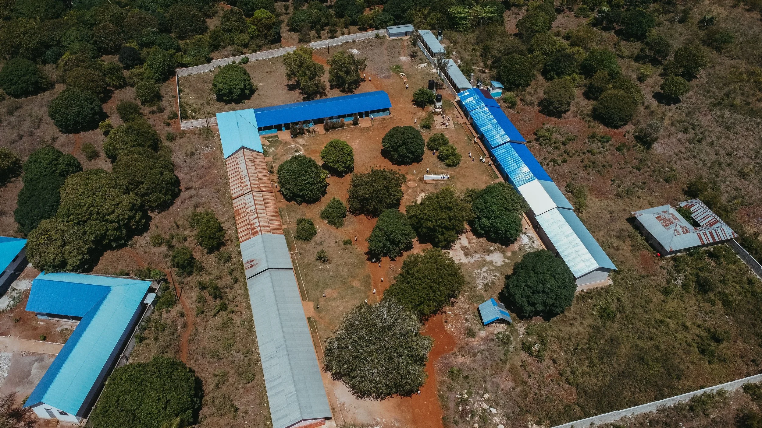 Aerial view of a school compound with blue-roofed buildings and trees, surrounded by a wall, in a rural area.