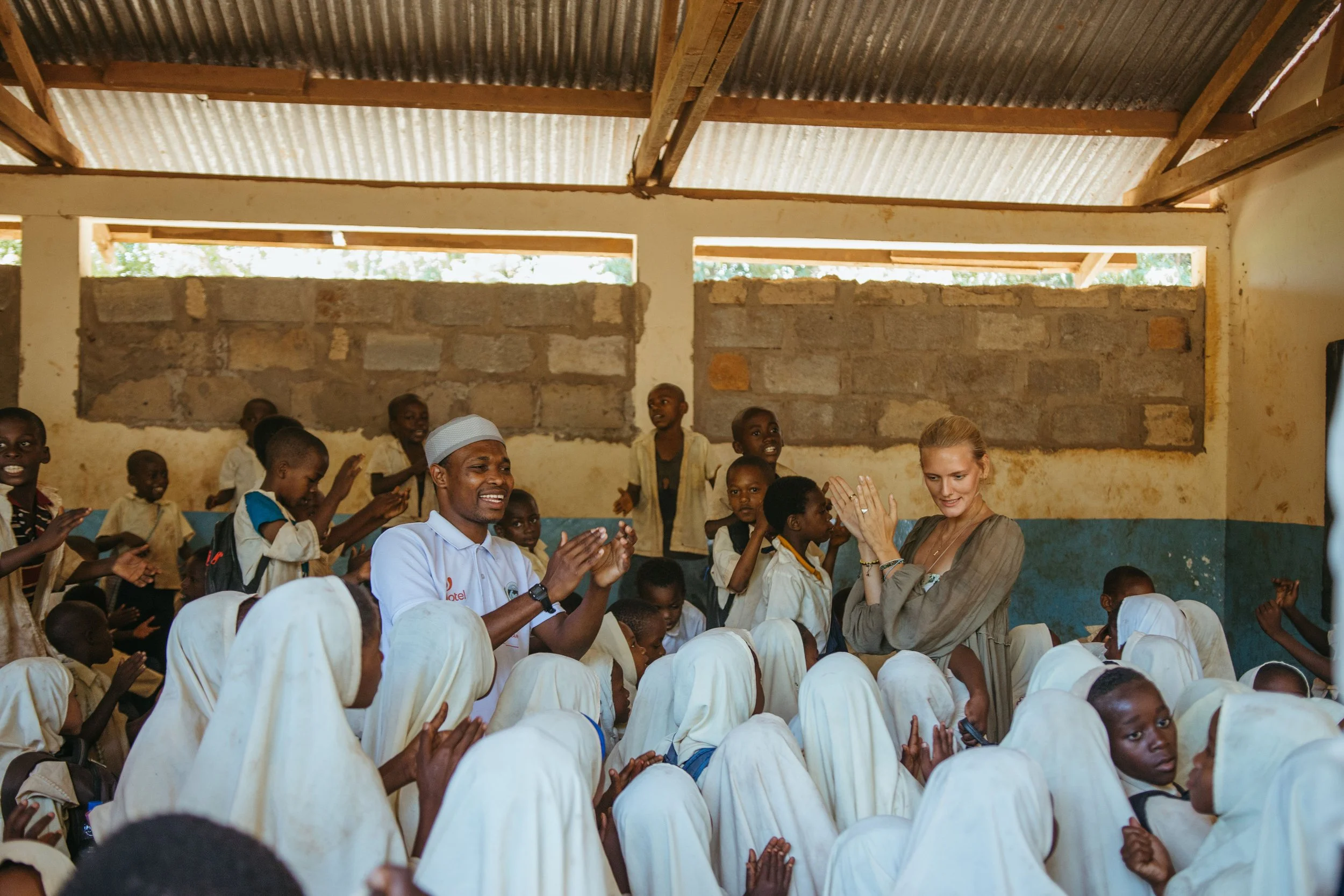 A group of children in beige uniforms sitting on the floor in a classroom, clapping and smiling, with two adults engaging with them, inside a room with unfinished brick walls and a corrugated metal roof.