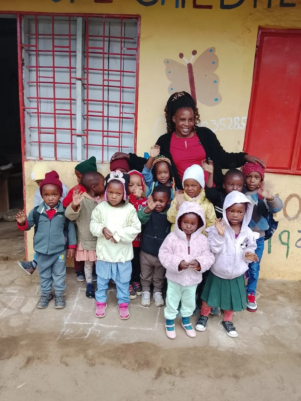 Group of young children with a woman, standing outdoors in front of a yellow wall with a butterfly painted on it. The children are waving and wearing colorful clothing and hats.