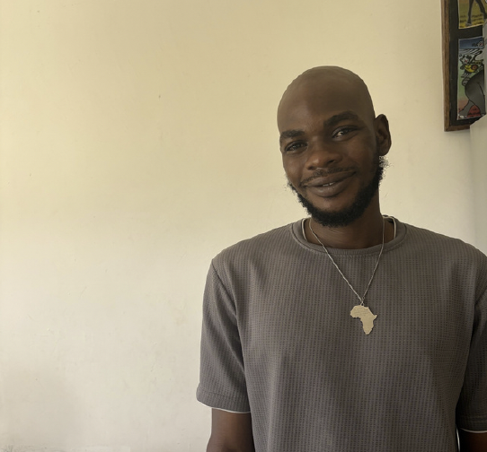 A smiling man with a beard and shaved head wearing a gray shirt and a necklace with an Africa-shaped pendant, standing indoors against a plain wall.