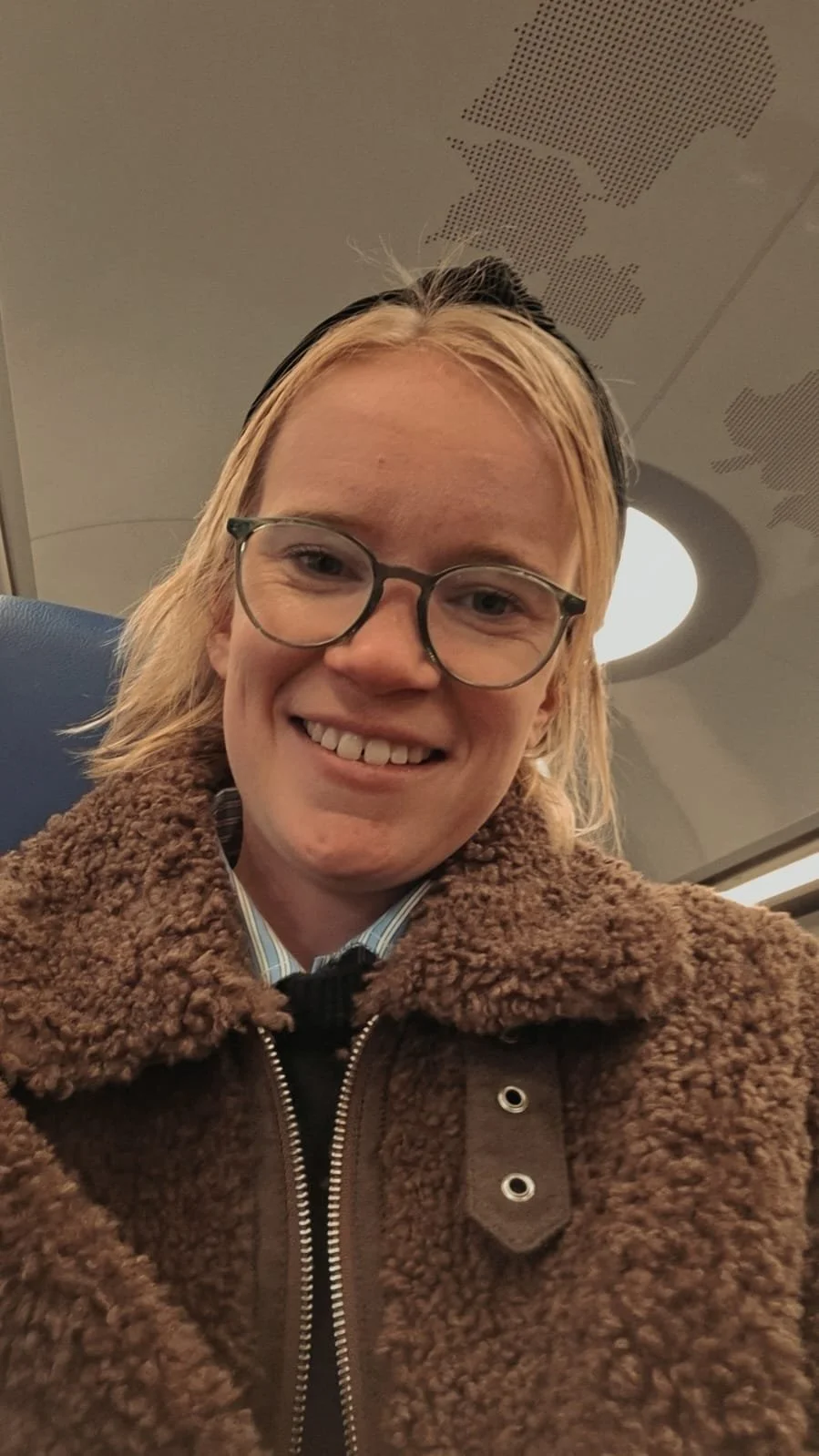 Smiling person with glasses, blonde hair, wearing a brown fuzzy jacket, on an airplane.