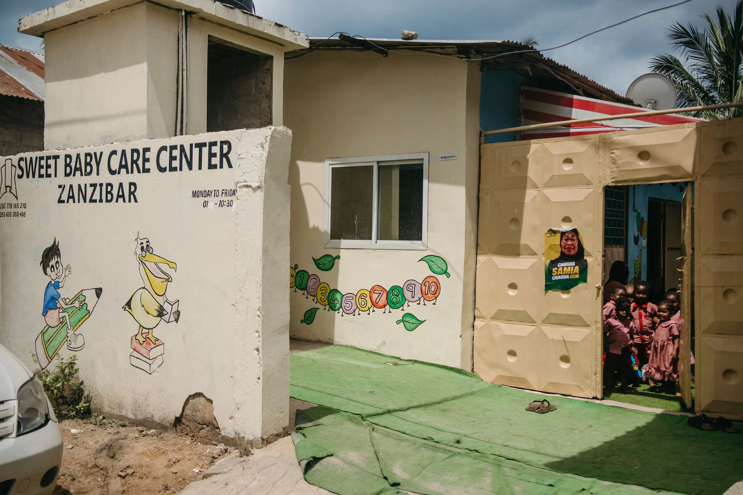 The image shows the entrance of a building with a sign that reads 'Sweet Baby Care Center Zanzibar,' with cartoon illustrations of children and a duck on the wall. There are children standing near the entrance, and the building has a satellite dish on the roof. The area outside the building has a mixture of paving and dirt, with some greenery.