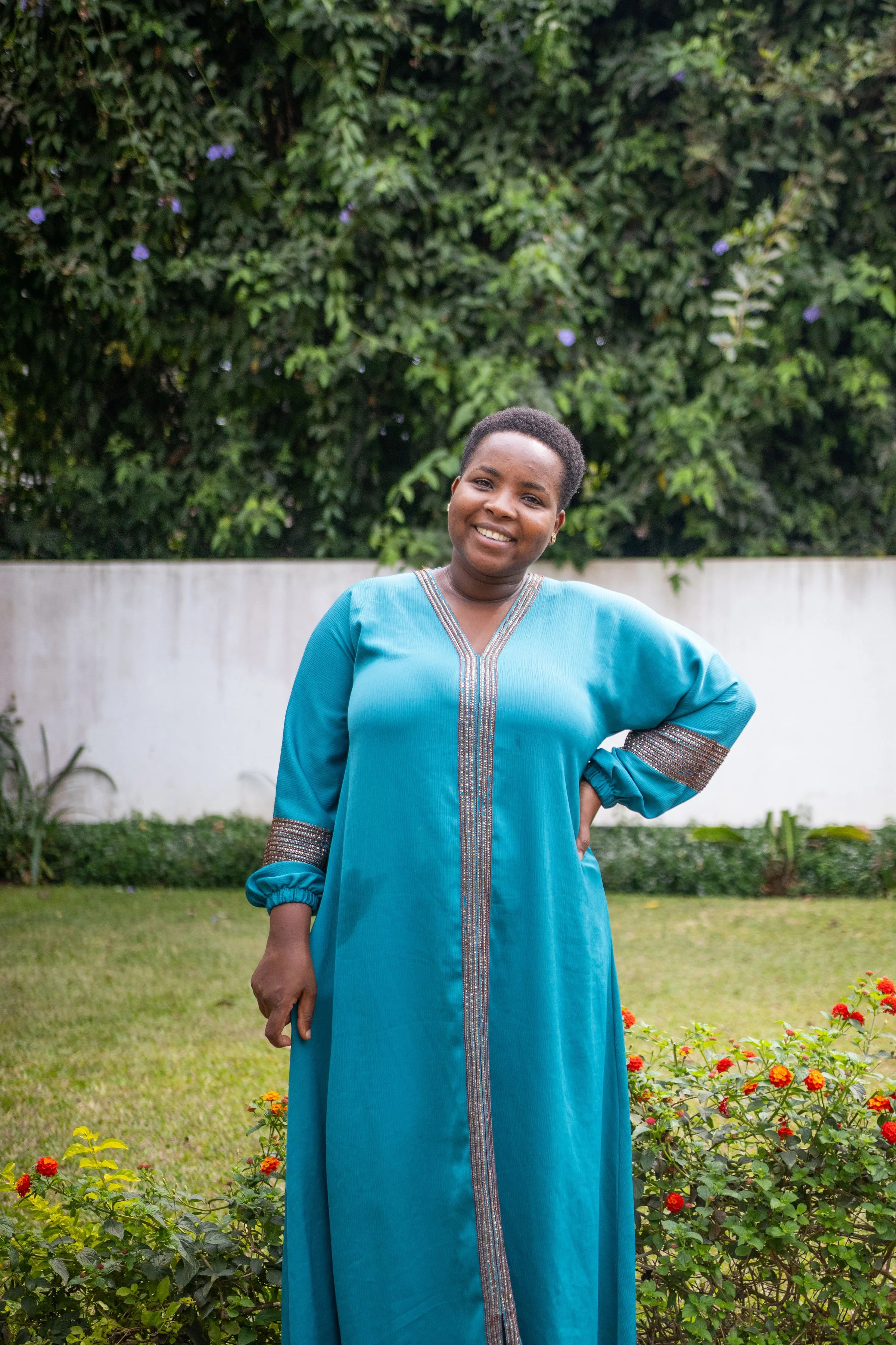 A woman standing outdoors in a garden with green foliage and colorful flowers, wearing a blue dress with silver embroidery and smiling at the camera.