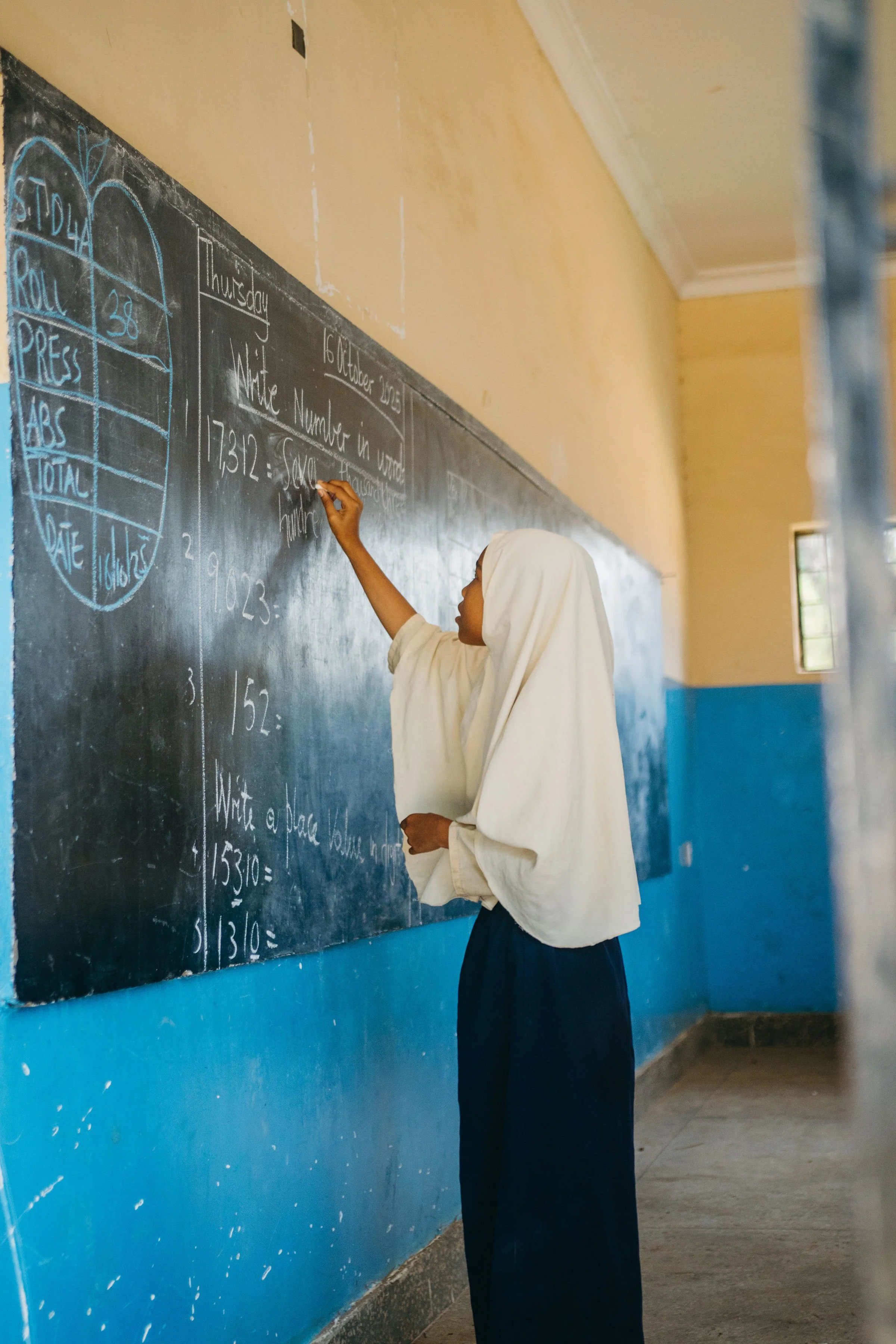 A girl in a white hijab and dark skirt writes on a black chalkboard in a classroom with yellow walls and a window.