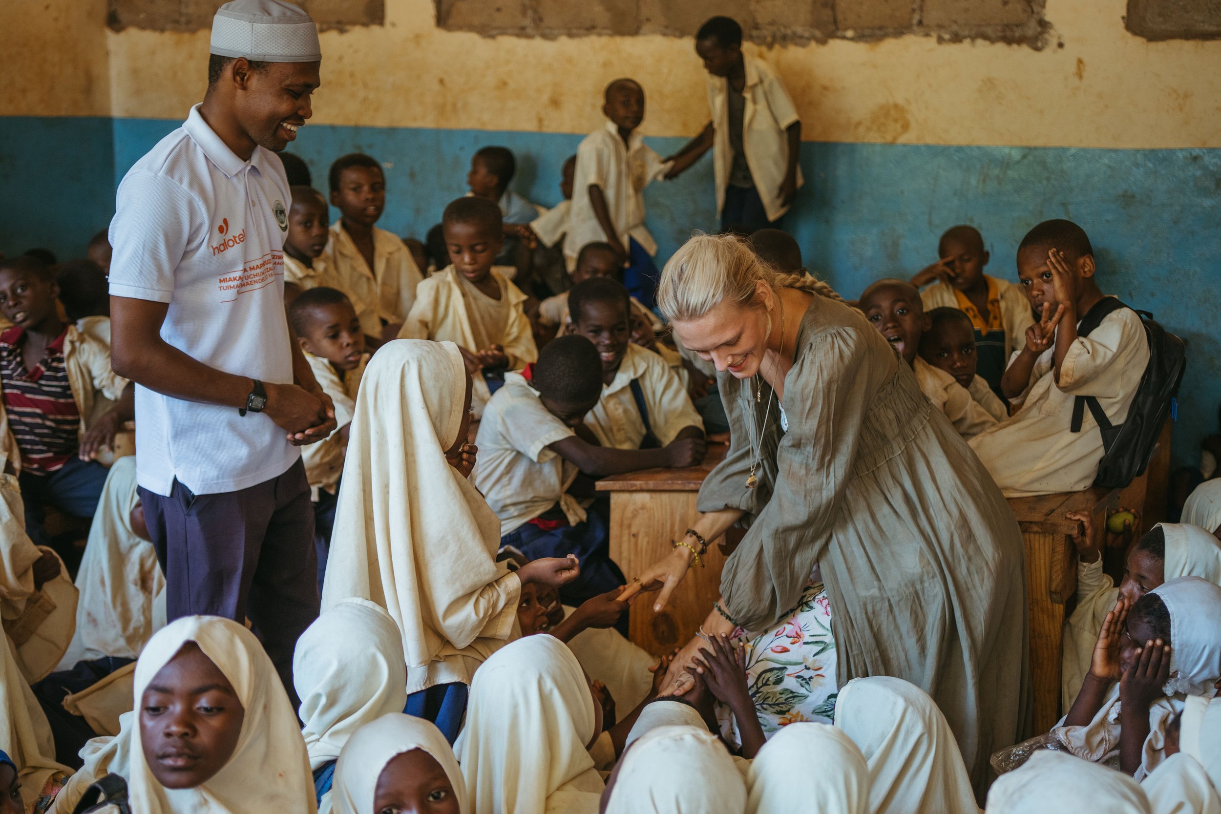 A group of children and women in a classroom, with a woman with blonde hair and a grey dress shaking hands with a girl wearing a white hijab. Other children are seated and standing, watching the interaction. The classroom has a worn yellow and blue wall.