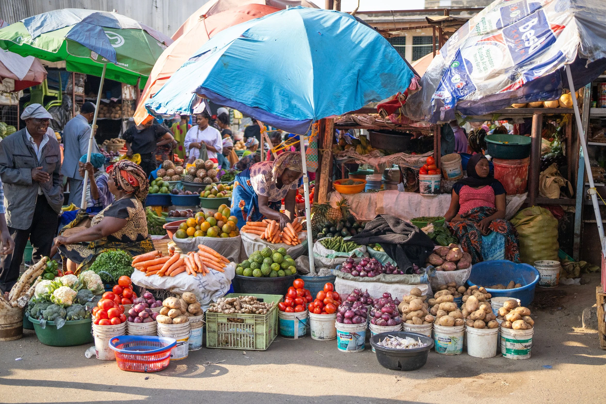 A bustling outdoor market with vendors and customers surrounded by stalls full of fresh vegetables, including carrots, tomatoes, potatoes, and green produce. Colorful umbrellas shade the market area.