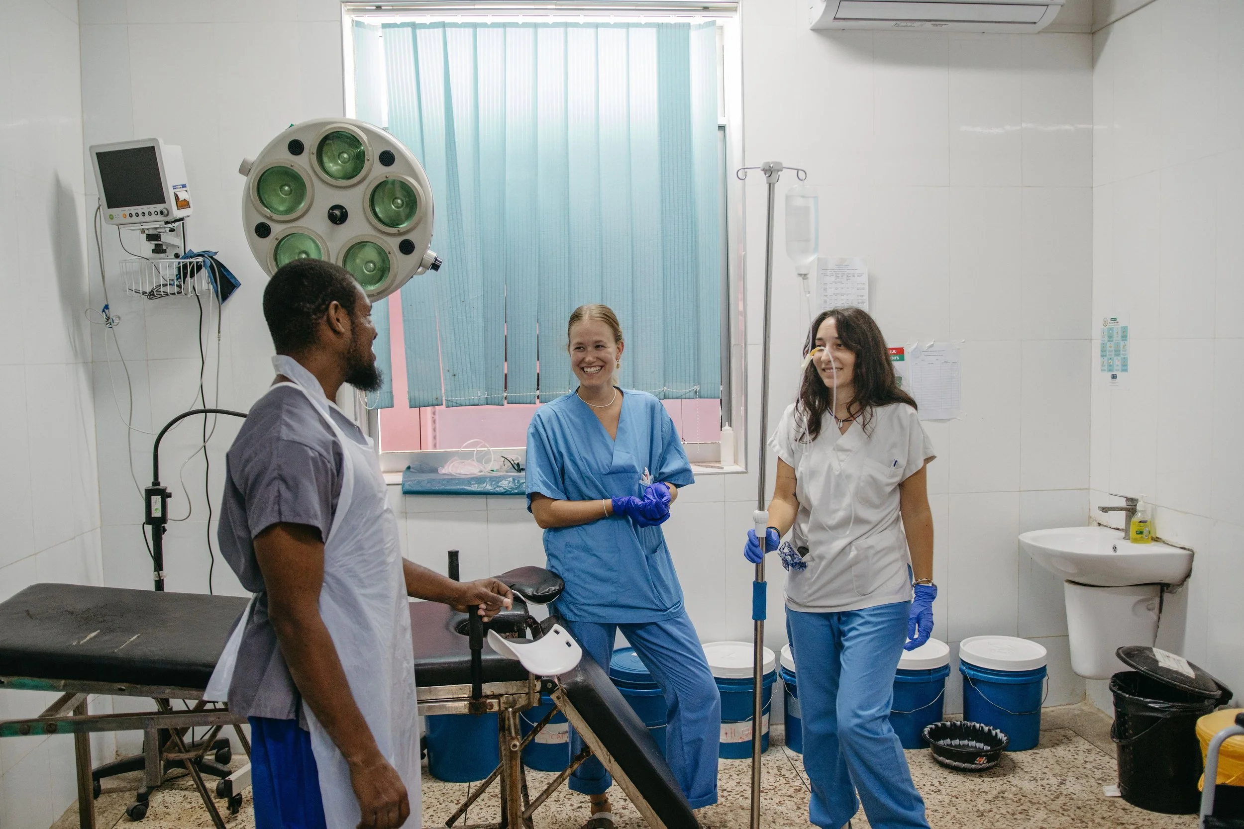 Three medical professionals in scrubs and gloves standing in a hospital room, smiling and talking with each other. One is seated on a hospital bed, another is holding an IV pole, and the third is standing near a sink. Medical equipment and blue bins are visible in the background.