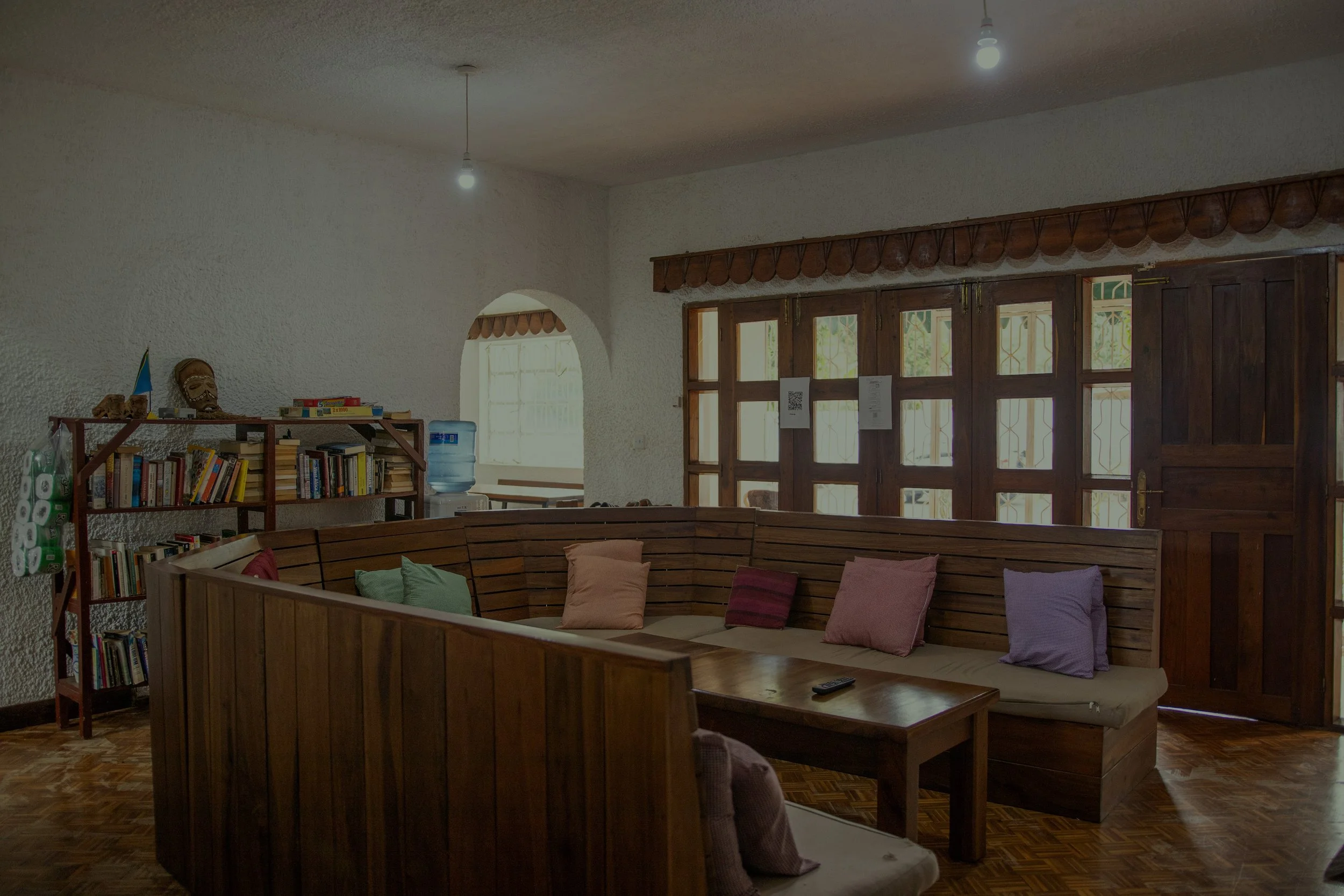 A cozy living room with a wooden corner sofa with colorful pillows, a bookshelf filled with books, and large windows with wooden frames allowing natural light, in a house with white textured walls and a wooden floor.
