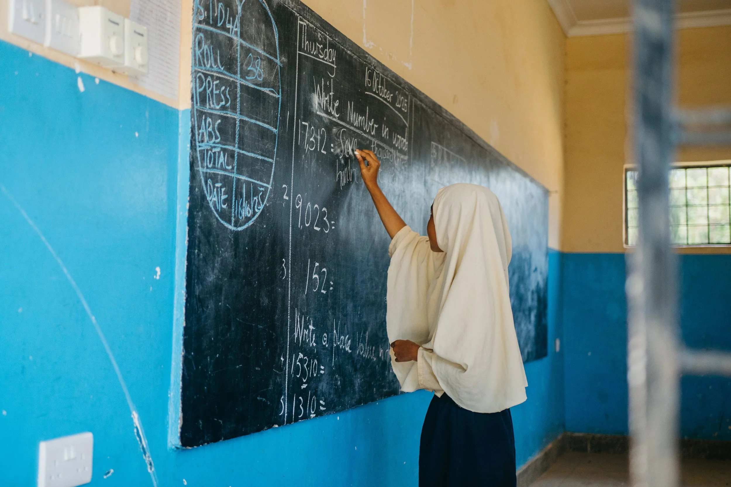 A student in a beige hijab writing on a black chalkboard in a classroom with yellow walls.