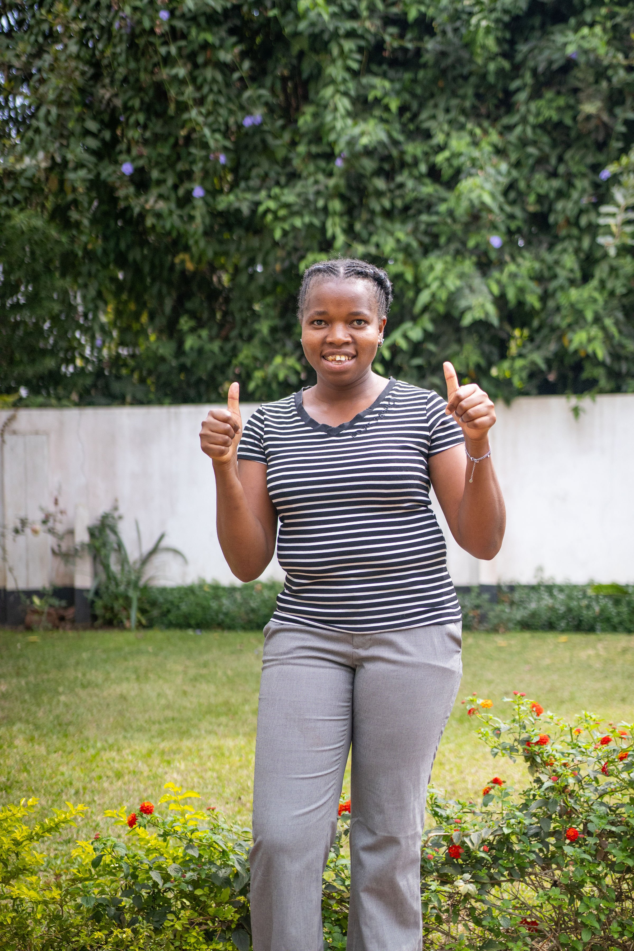 A woman standing in a garden with a white wall in the background, wearing a black and white striped shirt and gray pants, giving a thumbs-up with both hands and smiling.