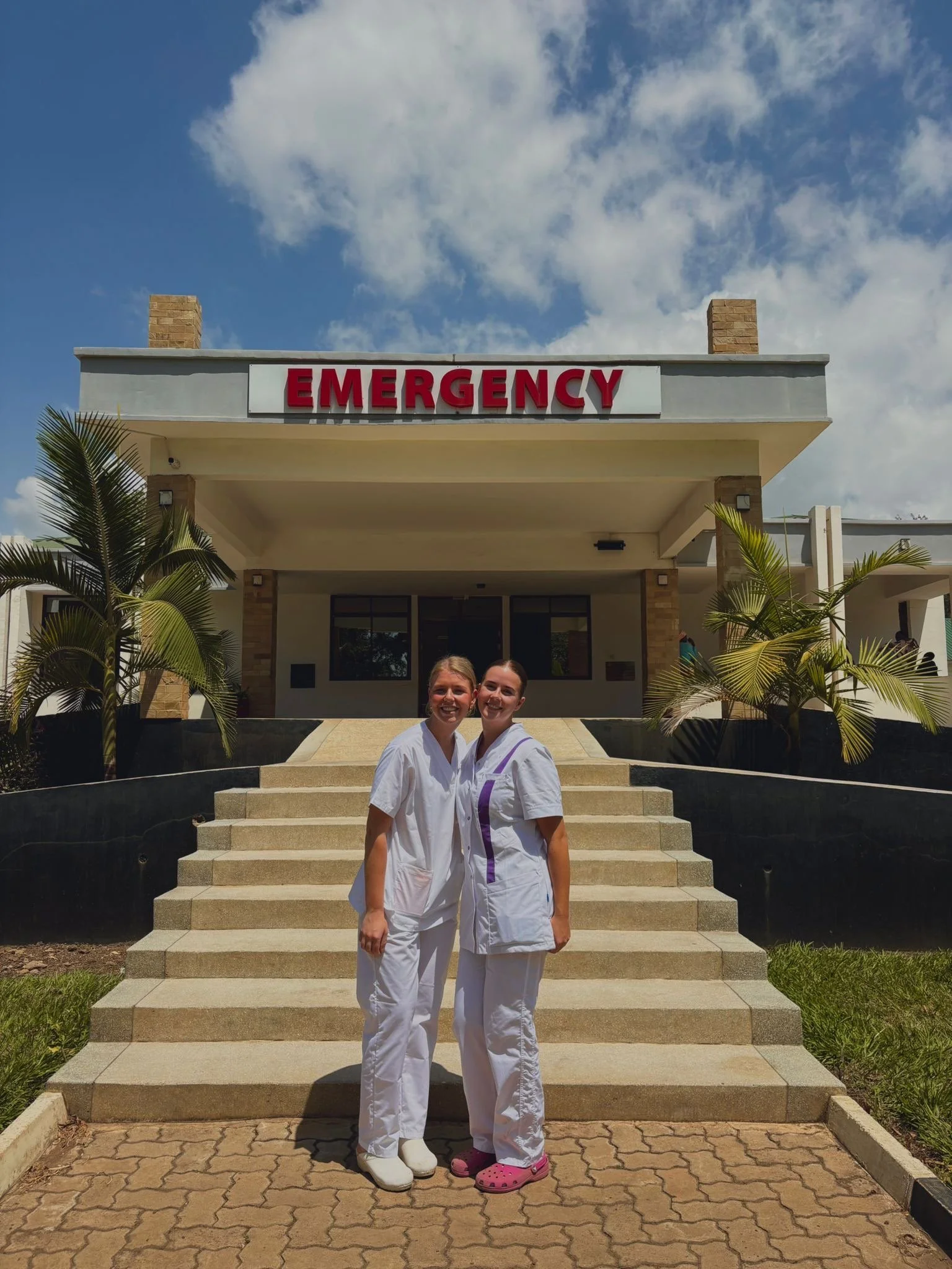 Two healthcare workers in white uniforms standing on outdoor steps in front of a building with a sign that reads 'EMERGENCY'. They are smiling, with palm trees on either side, under a partly cloudy sky.