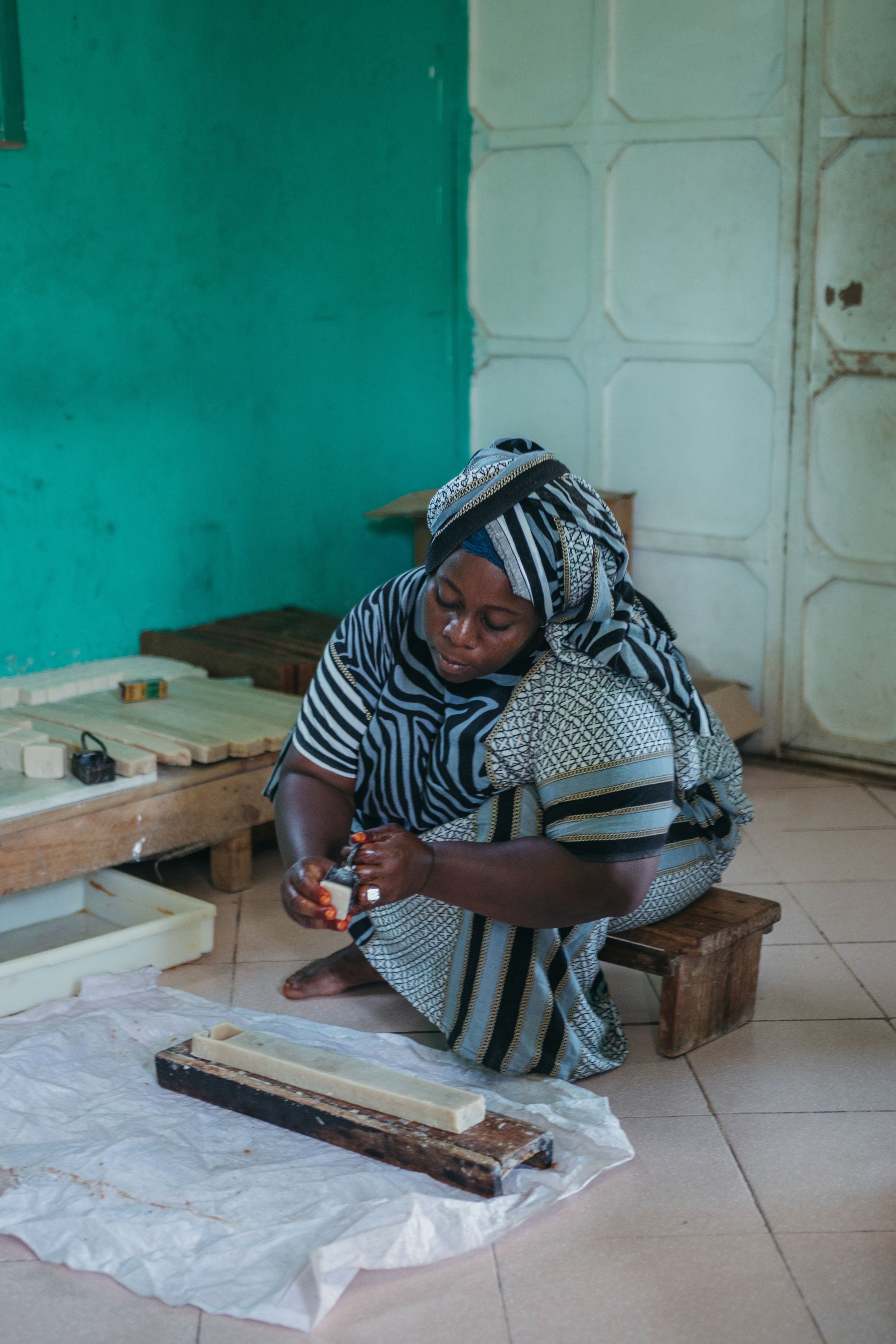 A woman dressed in traditional African attire is sitting on a small wooden stool, working with soap-making materials in a room with green and white walls. She appears focused as she handles a block of soap.