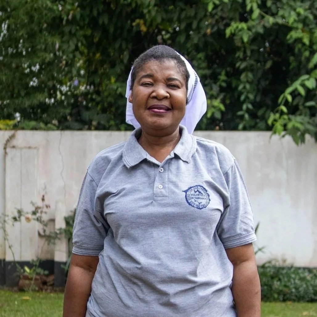A woman wearing a gray polo shirt with a logo on the chest and a white headscarf standing outdoors in front of a white wall and green trees.