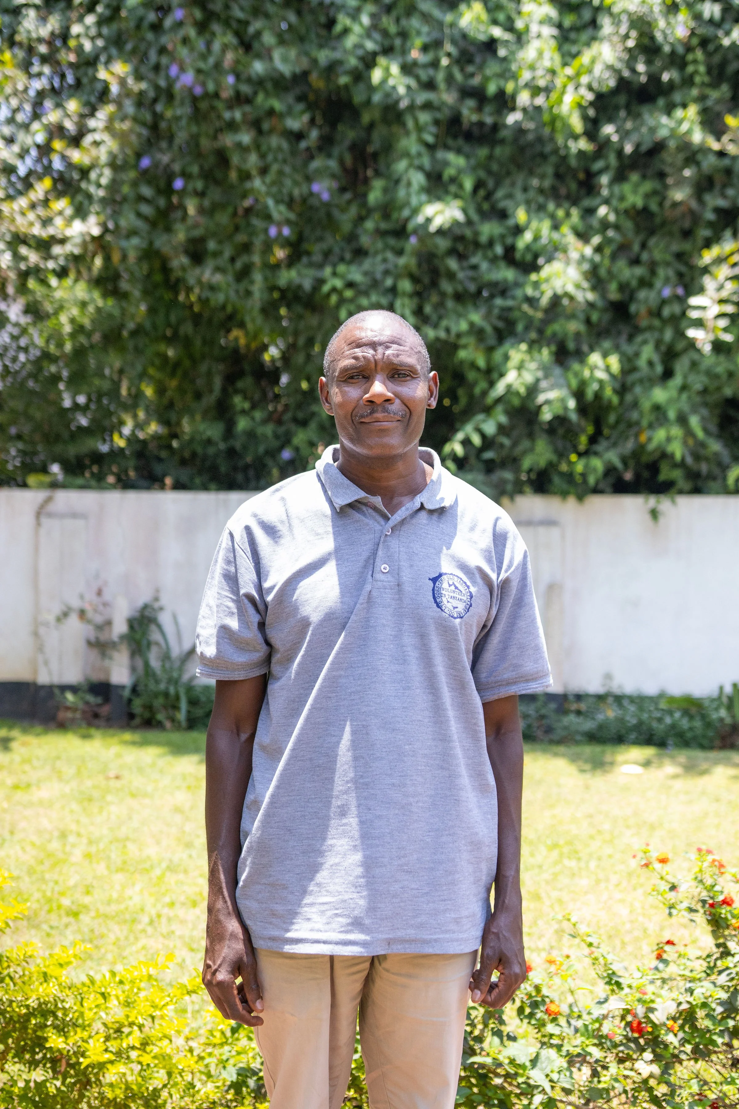 A man standing outside in front of a lush green leafy background with flowers at the bottom right. He is wearing a light grey polo shirt and beige pants, with a neutral facial expression.