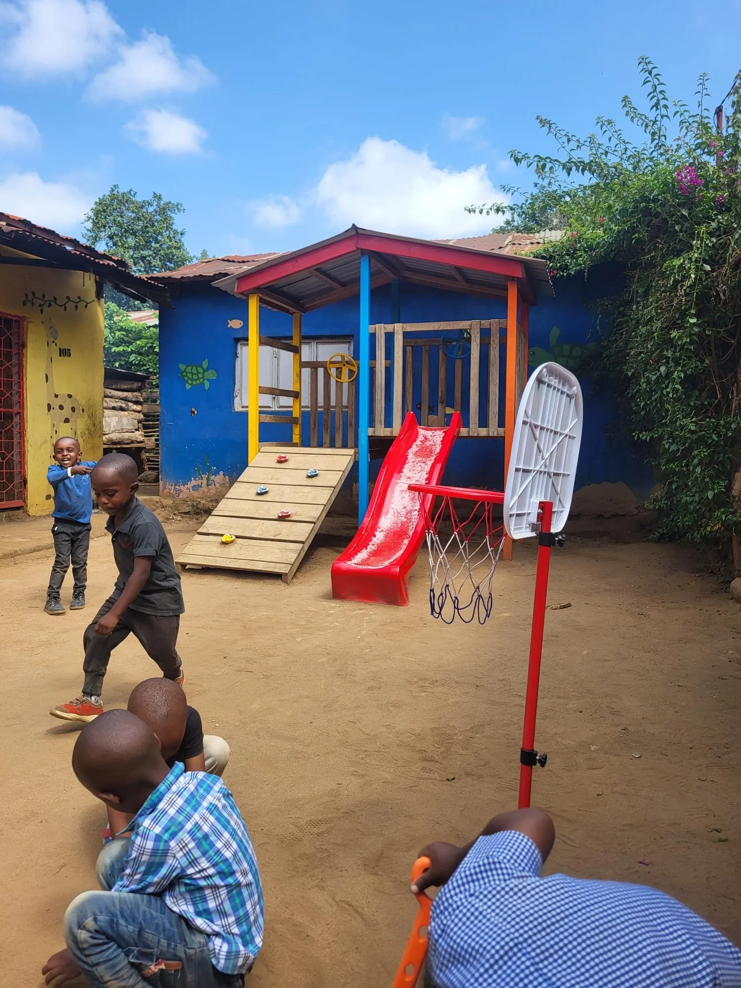 Children playing in a playground with slides, basketball hoop, and colorful painted walls in a sunny outdoor area.
