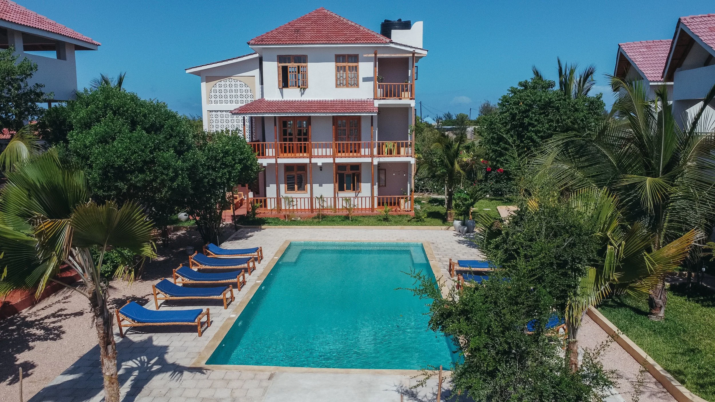 A white multi-story house with red-tile roof and wooden balconies, surrounded by lush greenery, with a rectangular swimming pool and sun loungers in the backyard.
