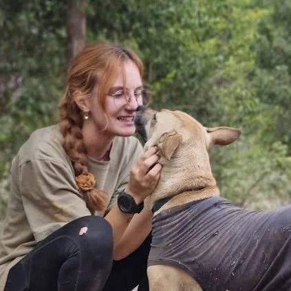 A woman with red hair and glasses smiling while holding and interacting with a large dog outdoors in a wooded area.