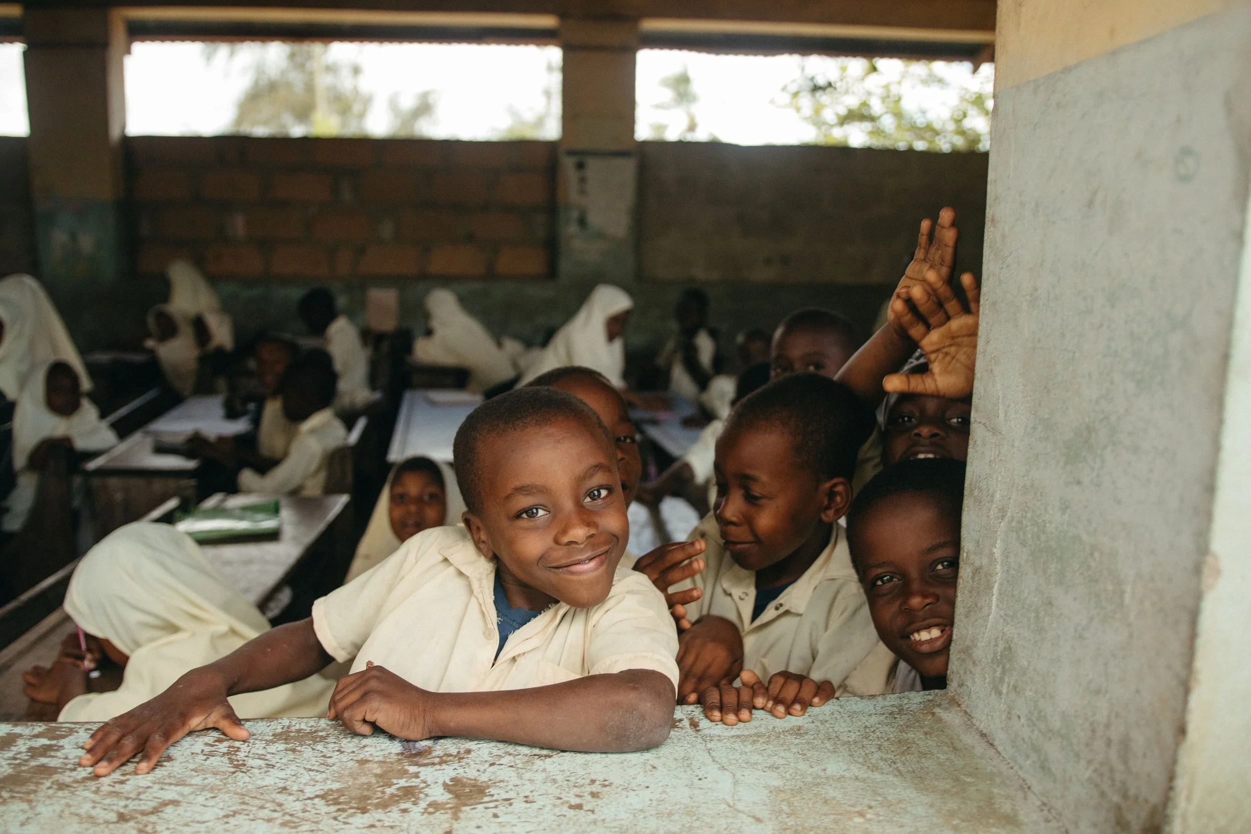 Smiling young children, wearing school uniforms, peering through a window in a classroom, with some children raising their hands, in a rural school setting.