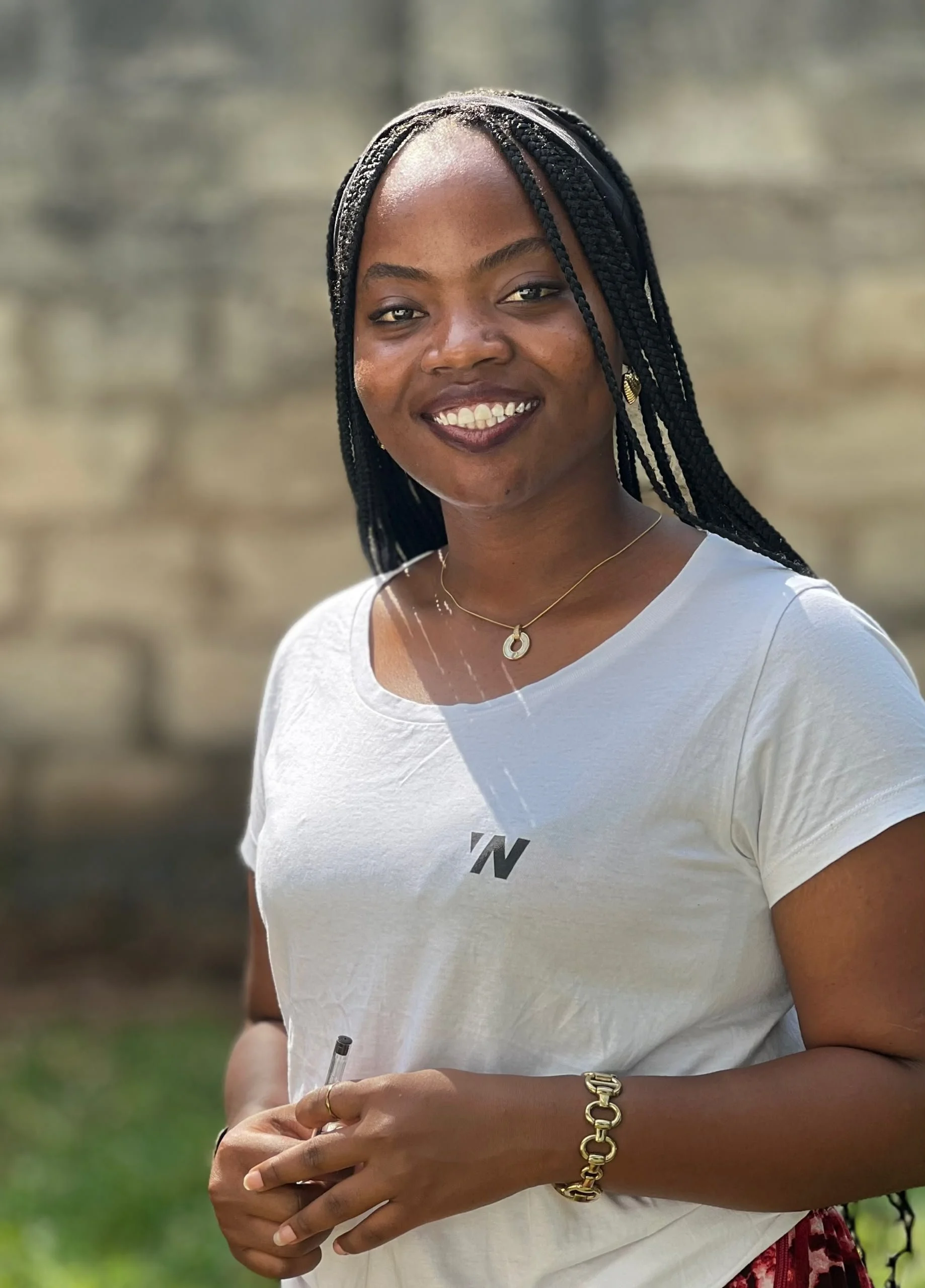 Close-up of a woman with braided hair smiling and holding a pen, wearing a white t-shirt and jewelry, outdoors with a blurred background.