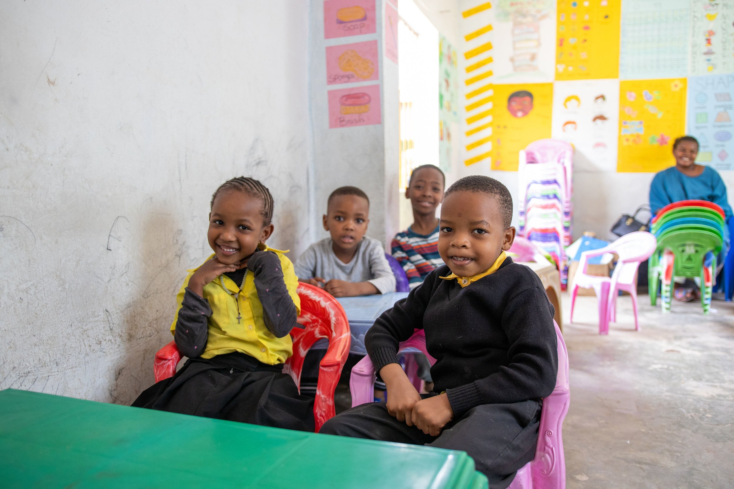 Group of smiling children sitting and standing around a table in a classroom, with colorful chairs and educational posters on the wall.