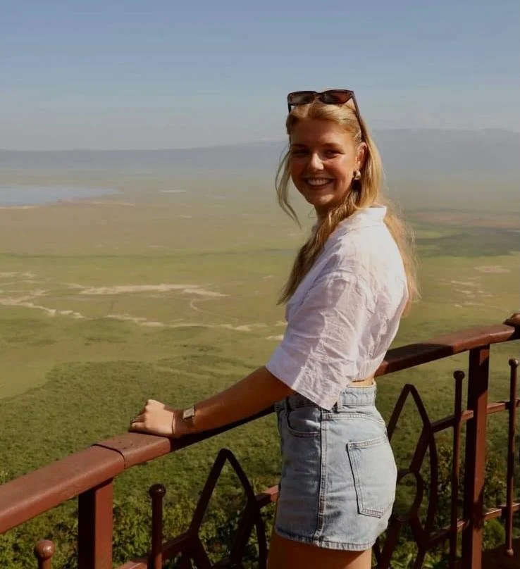 A young woman with blonde hair, sunglasses on her head, wearing a white top and denim shorts, smiling at the camera on a scenic outdoor balcony overlooking a vast green landscape and a distant body of water.