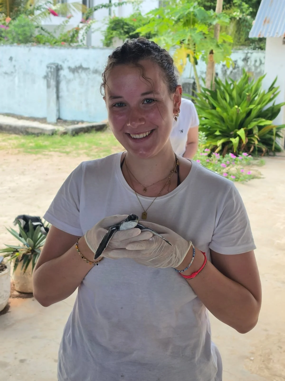 A young woman with curly hair and a white t-shirt is holding a small black and white snake with gloved hands, smiling at the camera outdoors surrounded by plants and a garden.