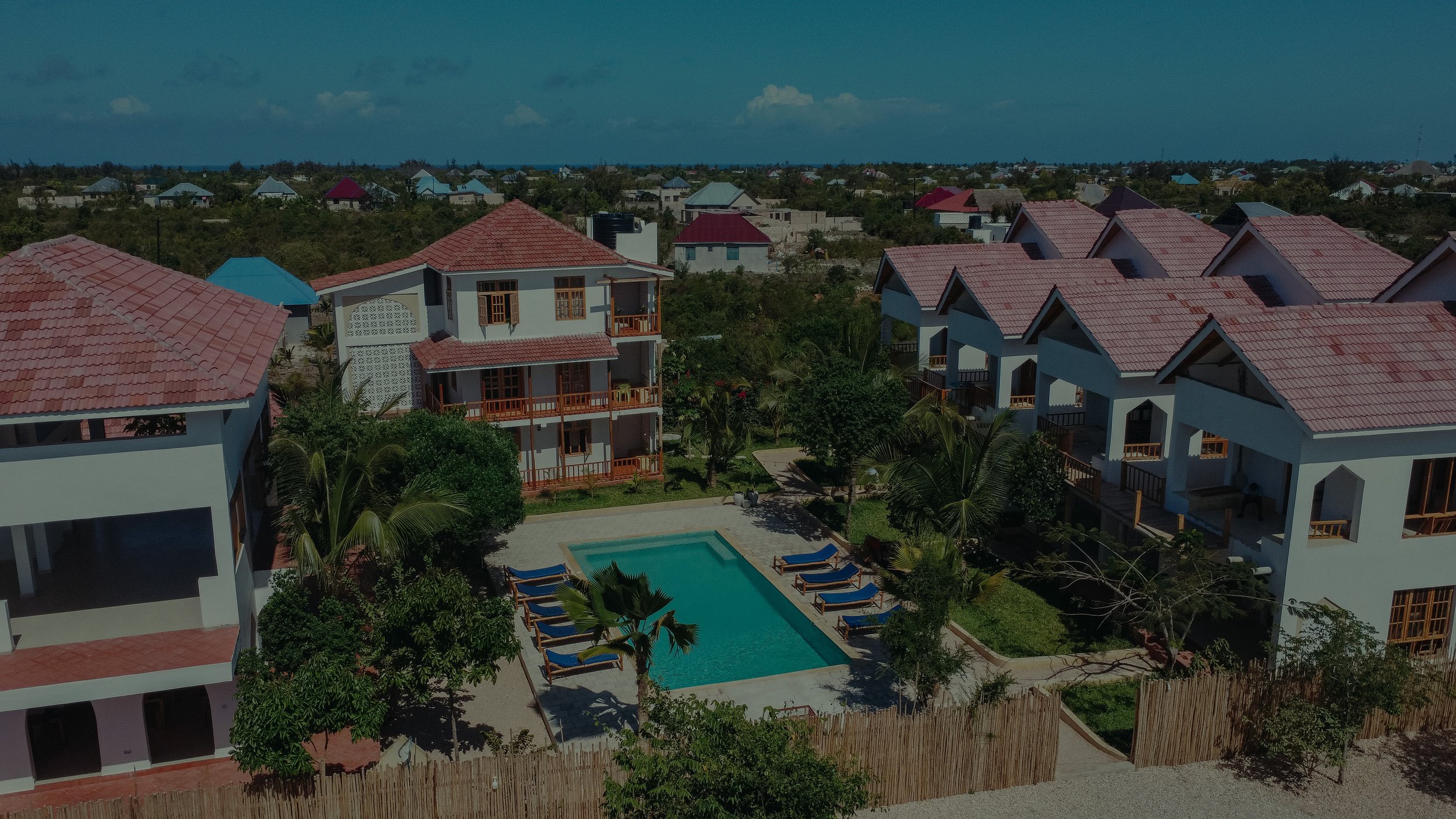 Aerial view of a resort with white buildings with red roofs, a swimming pool surrounded by lounge chairs, and lush green trees, set against a distant landscape of more houses and trees under a partly cloudy sky.