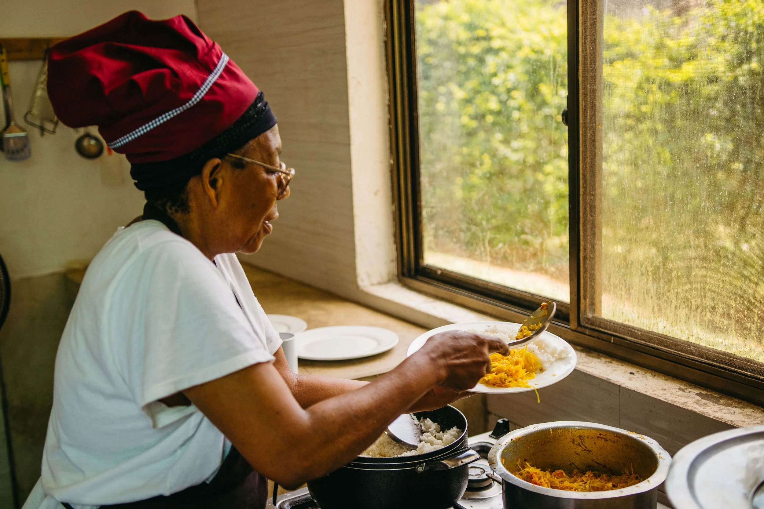 Woman with glasses and a headwrap serving herself food from large pots near a window with a view of trees.