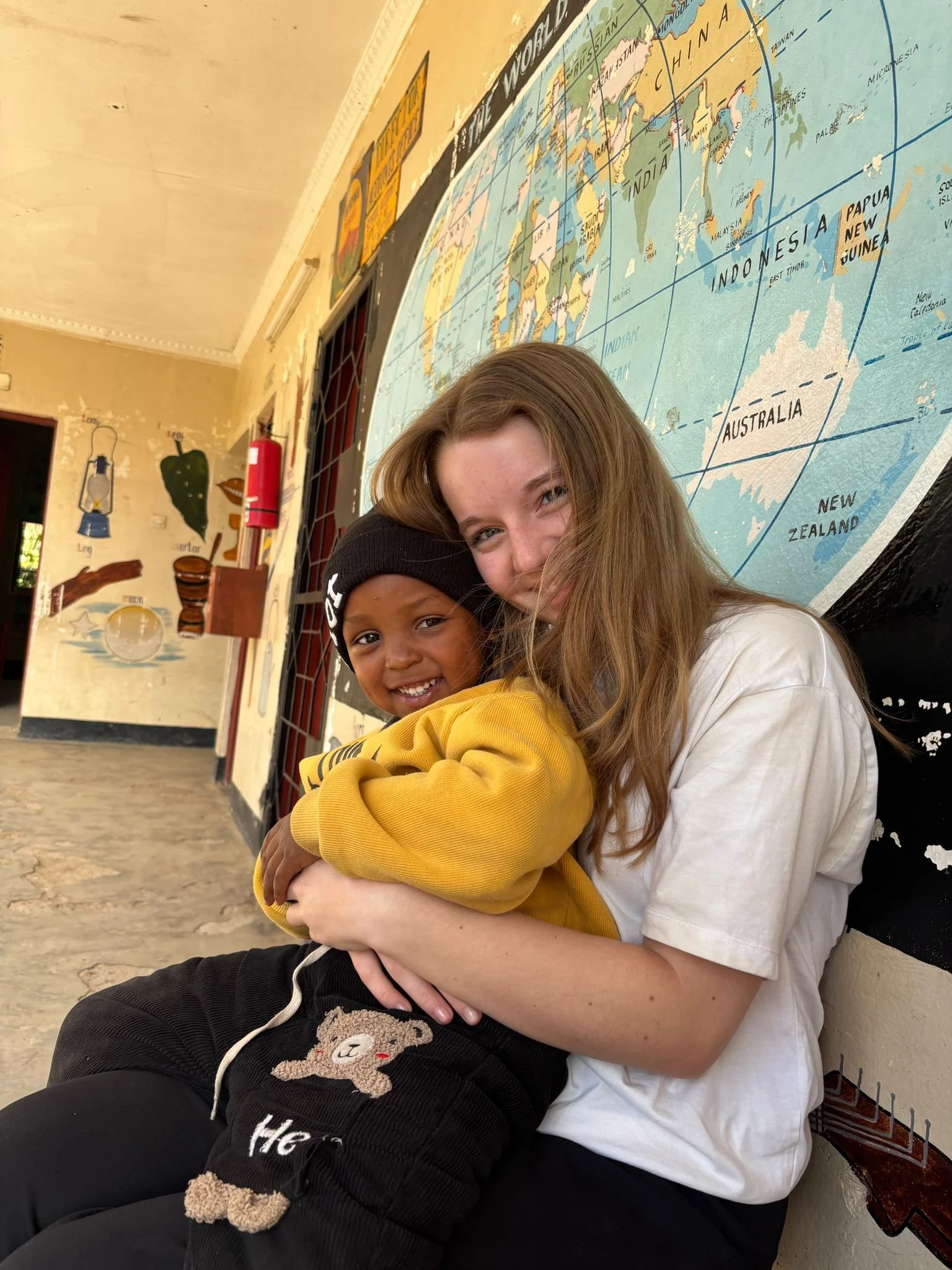 A young boy with dark skin wearing a black hoodie and black pants, and a young girl with light skin and long red hair in a white shirt, are hugging and smiling at the camera inside a classroom with a large world map on the wall behind them.